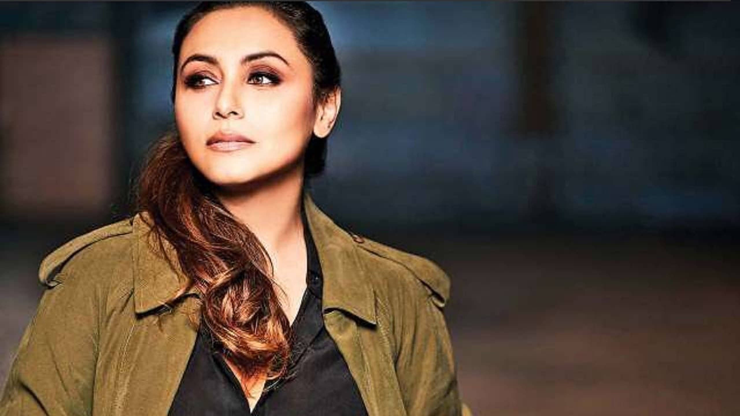 Rani Mukerji 桌布 壁紙 — 一位長棕髮女性穿著橄欖綠色外套，背景模糊昏暗。
