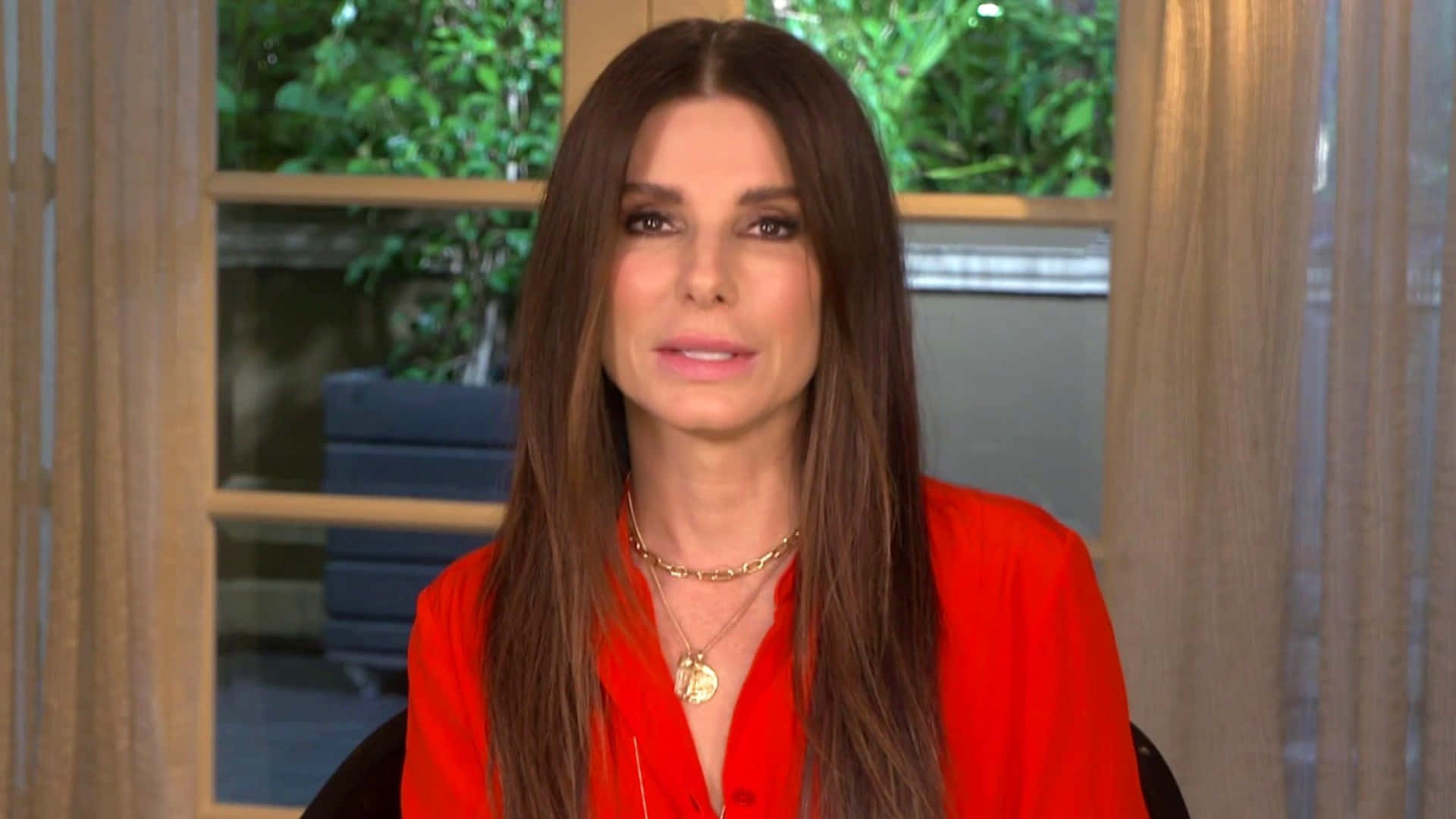 Sandra Bullock 2021 桌布 壁紙 — 一位長棕髮女性穿著紅色襯衫和金項鍊，坐在室內，身後窗戶可見綠色植物。