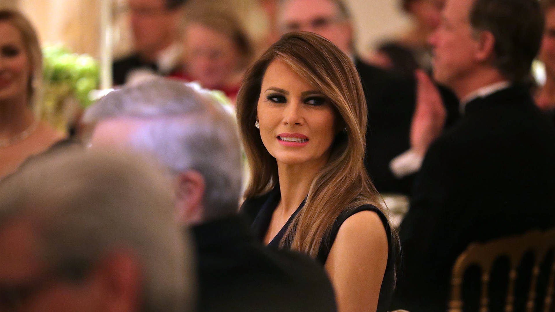 Melania 桌布 壁紙 — 一位長棕髮的女性在正式晚宴上微笑，周圍的賓客模糊不清。