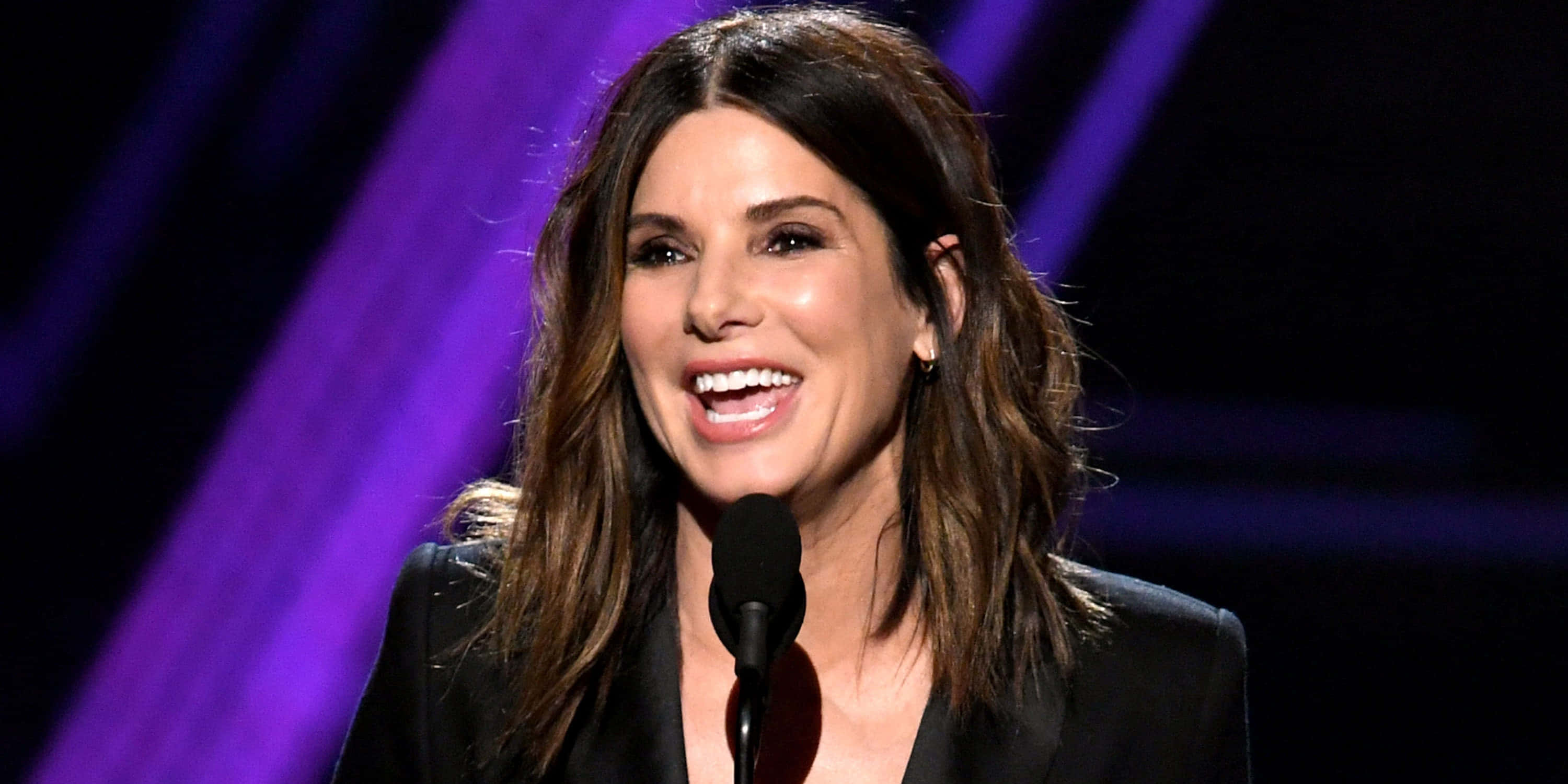 Sandra Bullock 2021 桌布 壁紙 — 一位長棕髮的女性在紫色燈光照耀的舞台上對著麥克風燦爛微笑說話。