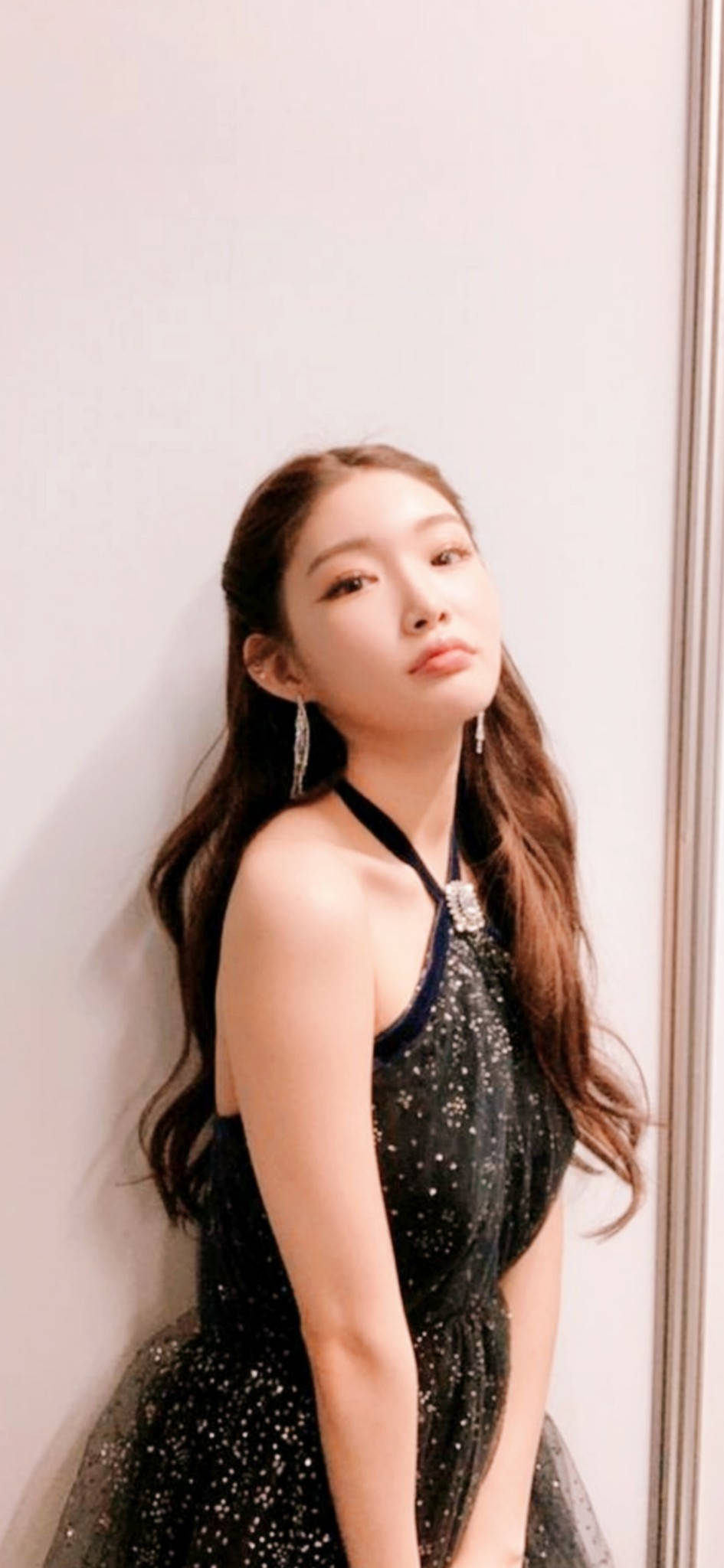 Chungha 桌布 壁紙 — 一位長棕髮的年輕女士靠著素牆，身穿閃亮黑色連衣裙和垂墜耳環。