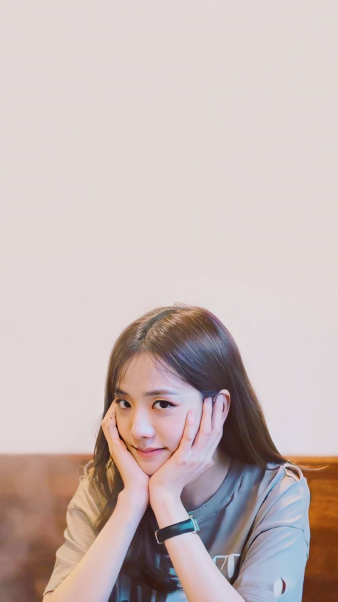 Jisoo Cute 桌布 壁紙 — 一位長棕髮的年輕女子將下巴擱在手上，對著純米色牆壁溫柔微笑。