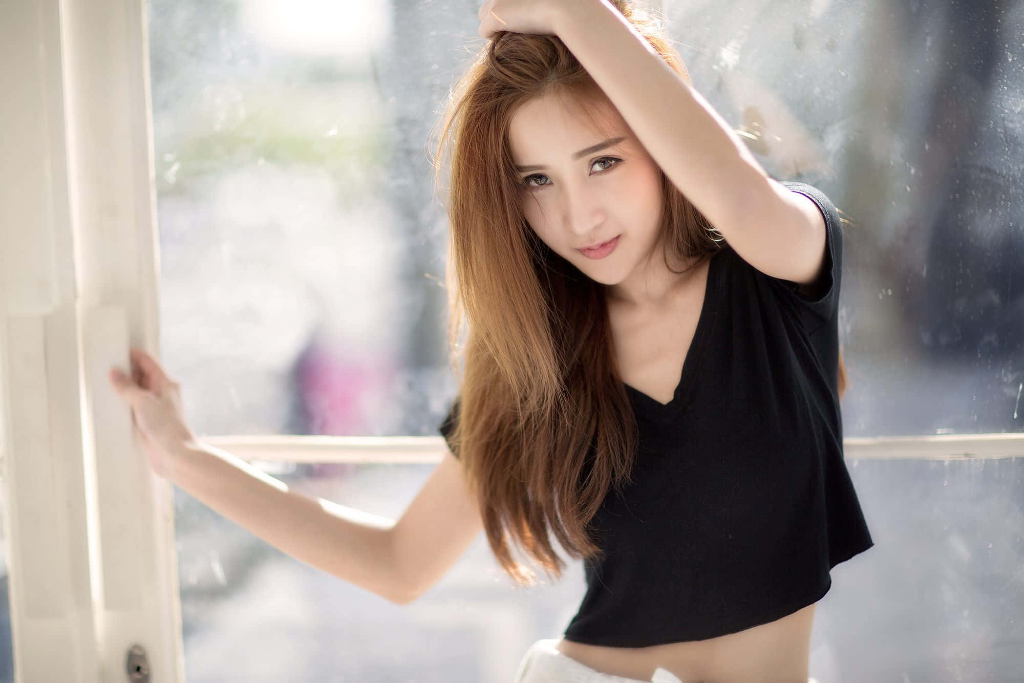美女 桌布 壁紙 — 一位長棕髮的年輕女性穿著黑色短版上衣，在柔和自然光中倚靠著雨窗。