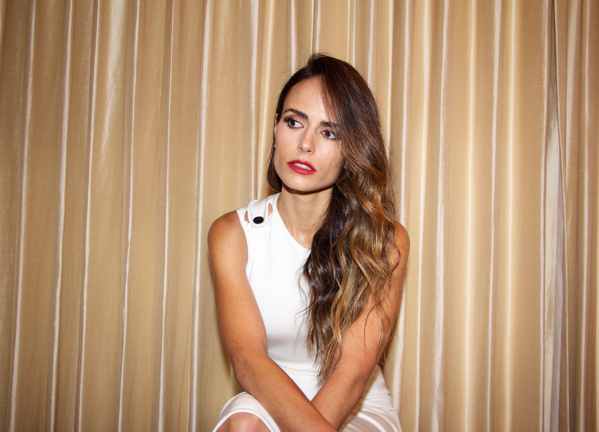 Jordana Brewster 桌布 壁紙 — 一位長波浪髮、紅唇的女性，身穿白色連衣裙，靠著米色窗簾坐著。