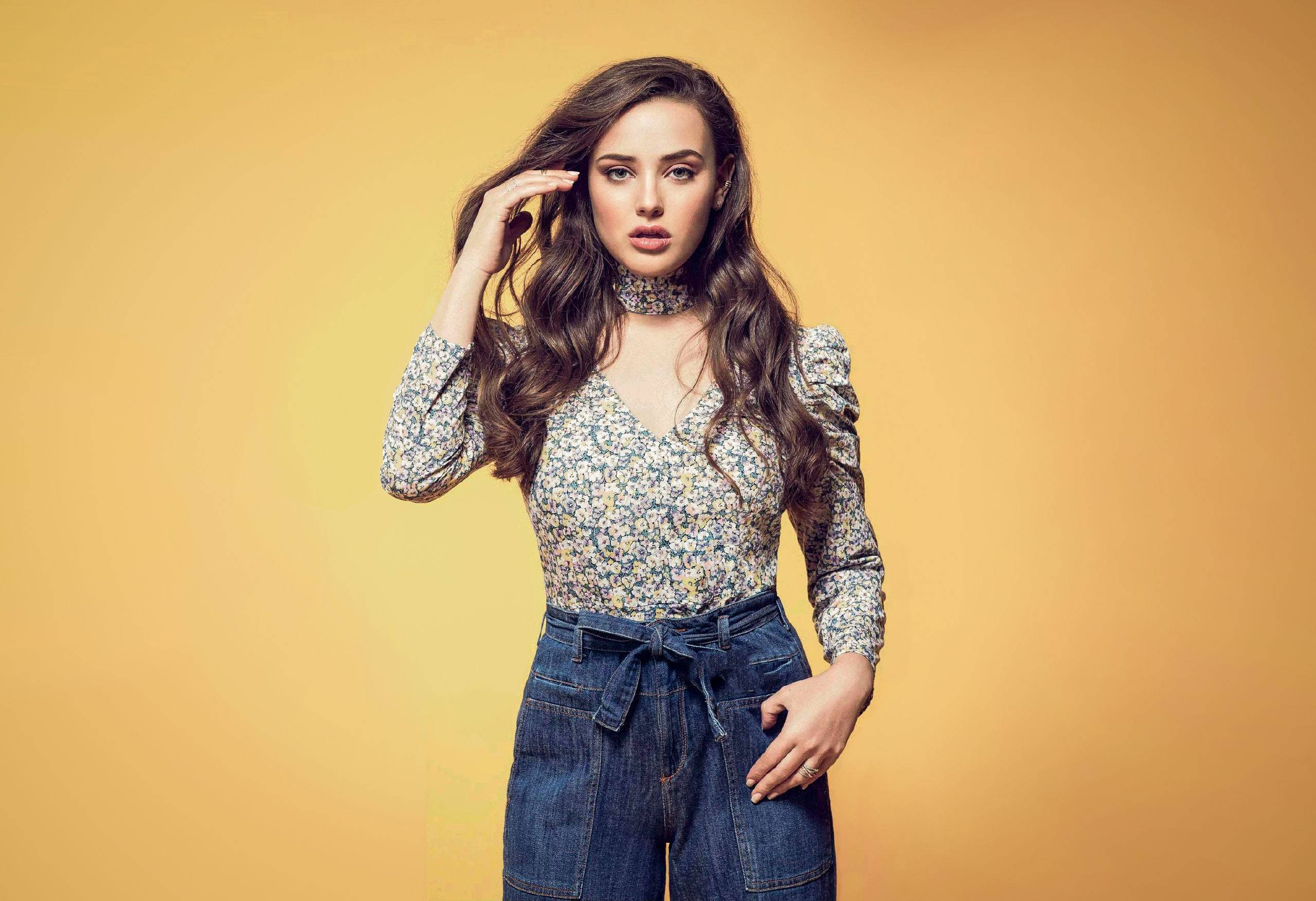 Katherine Langford 桌布 壁紙 — 一位長波浪髮的女性在溫暖的黃色背景前自信地擺姿勢，身穿花卉襯衫和牛仔褲。