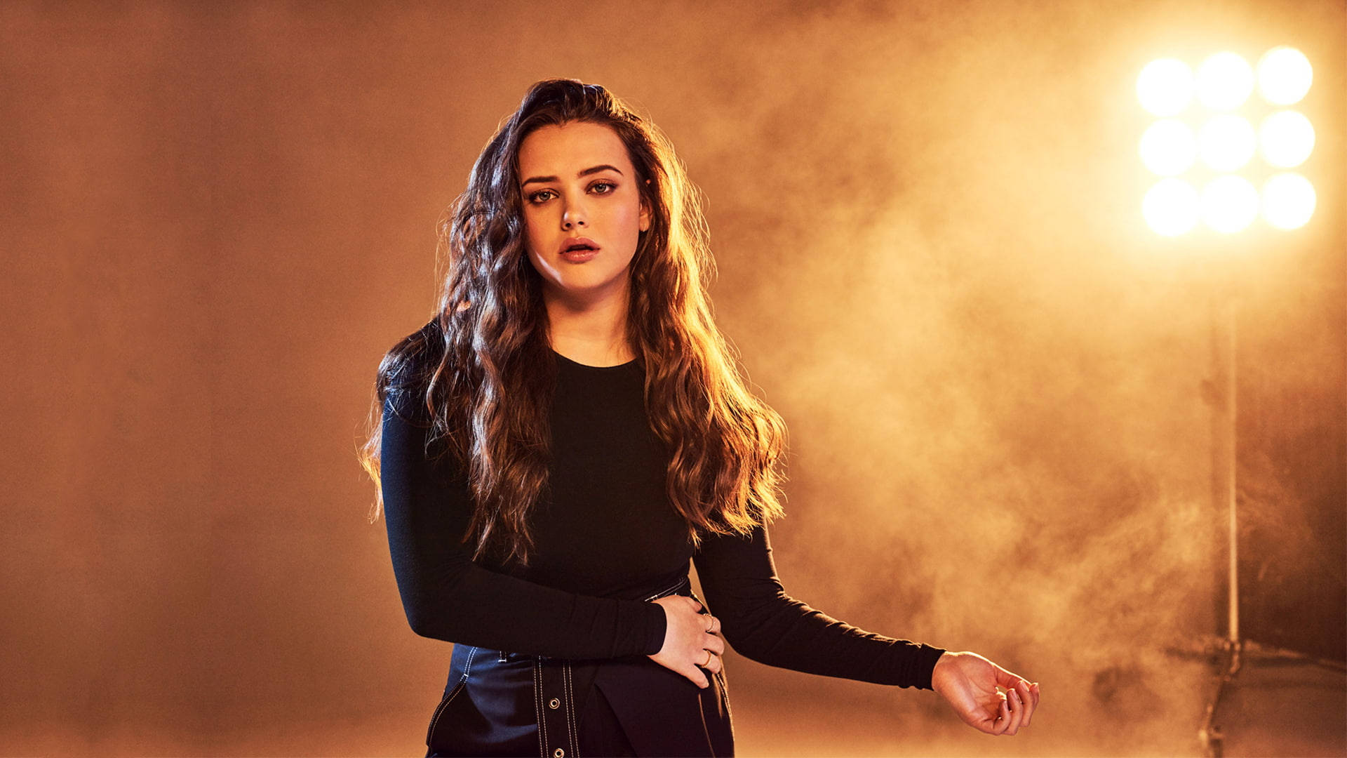 Katherine Langford 桌布 壁紙 — 一位長波浪髮的女性自信地站在溫暖的金色工作室燈光下，煙霧在她周圍旋轉。