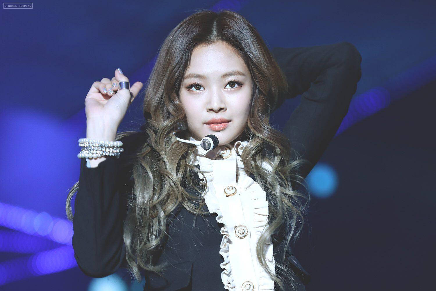 Jennie 桌布 壁紙 — 一位長波浪髮的女表演者在藍色舞台燈光下，穿著黑白服裝對著麥克風唱歌。