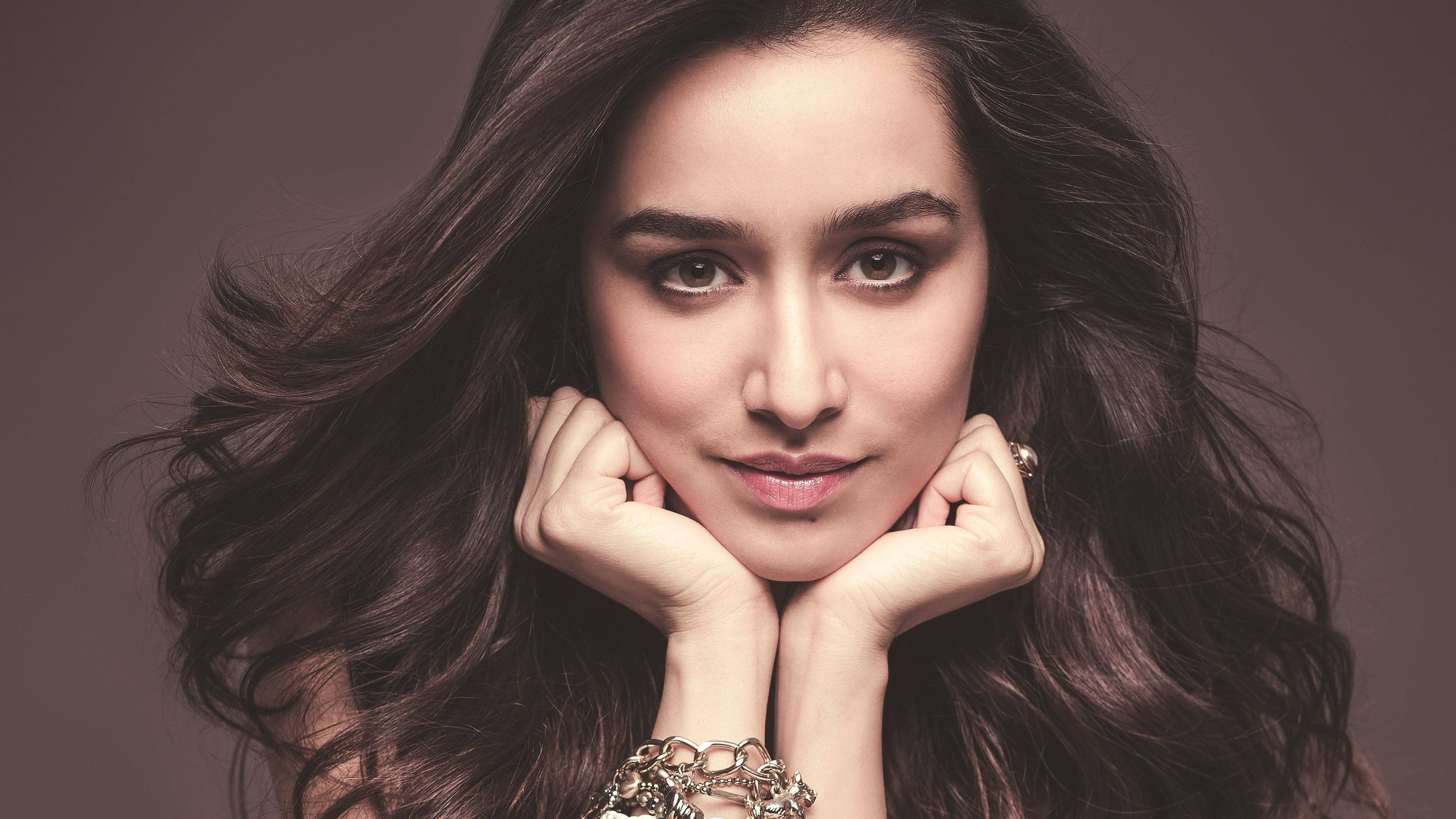 Shraddha Kapoor 桌布 壁紙 — 一位長深色波浪髮的女性將下巴擱在手上，背景為柔和的棕色。