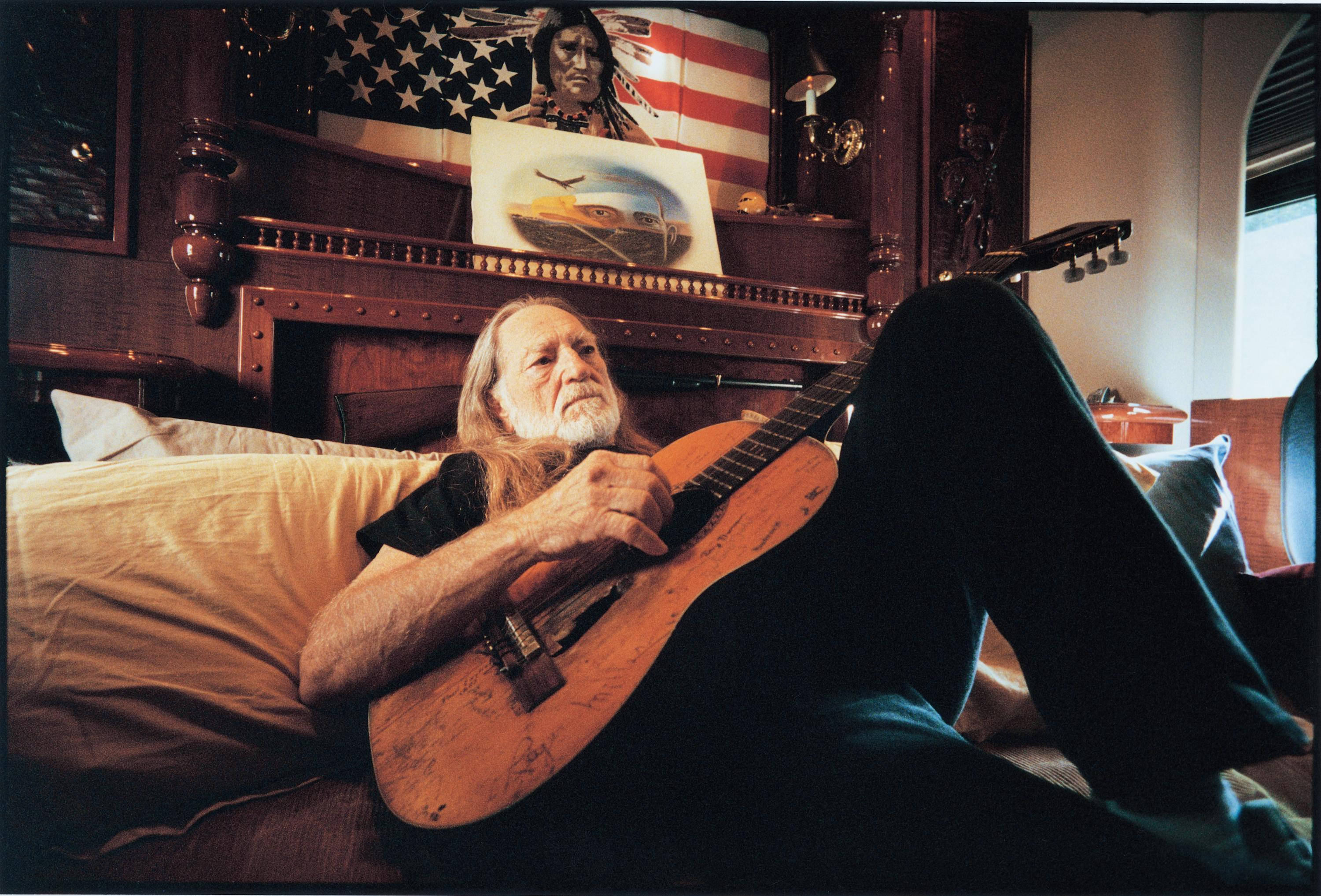 guitar player 桌布 壁紙 — 一位長白髮的長者躺在床上，手持木吉他，周圍是美式裝飾和溫暖的燈光。