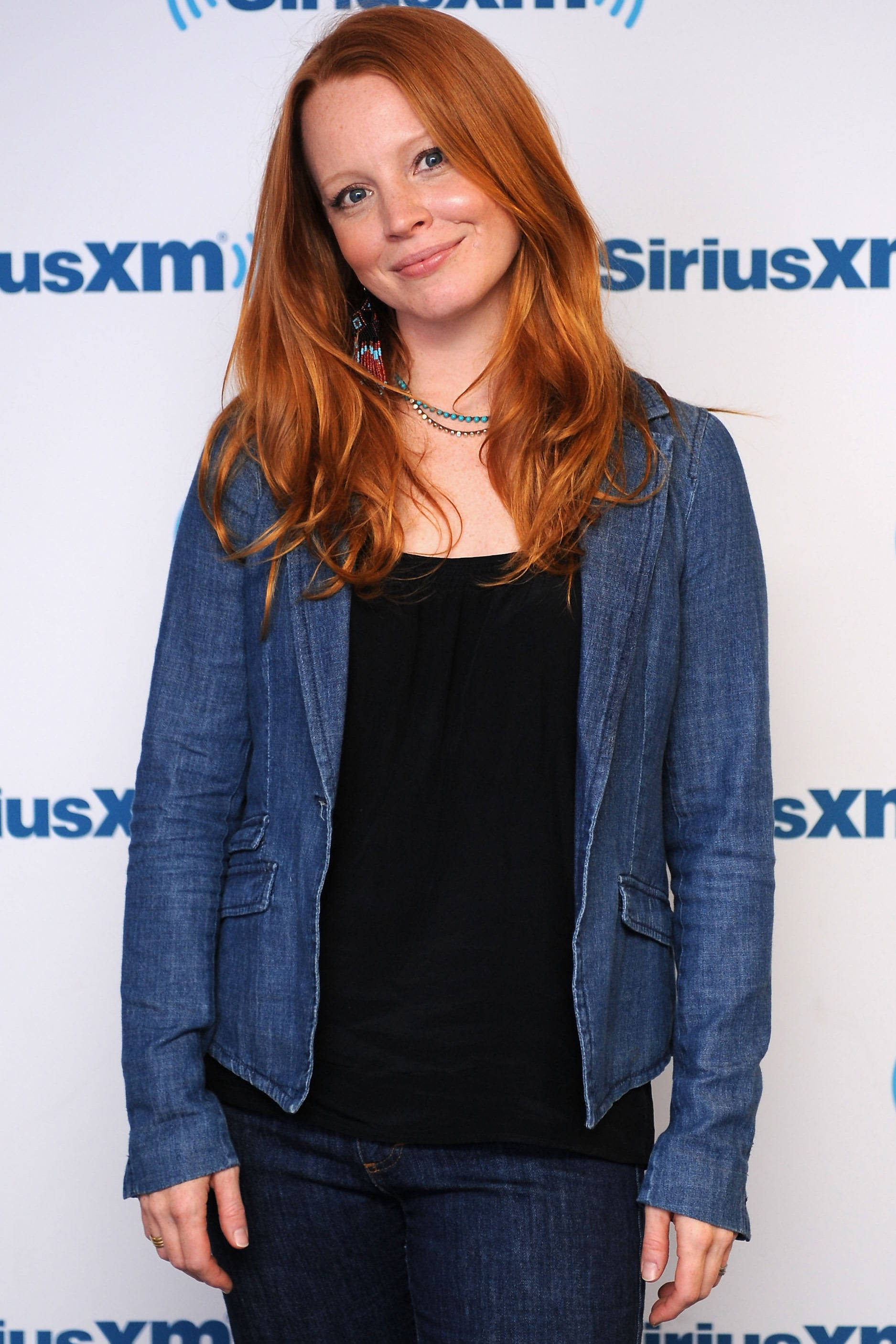 Broadway 桌布 壁紙 — 一位長紅髮的女性穿著牛仔外套和牛仔褲，在SiriusXM品牌背景前溫暖地微笑。