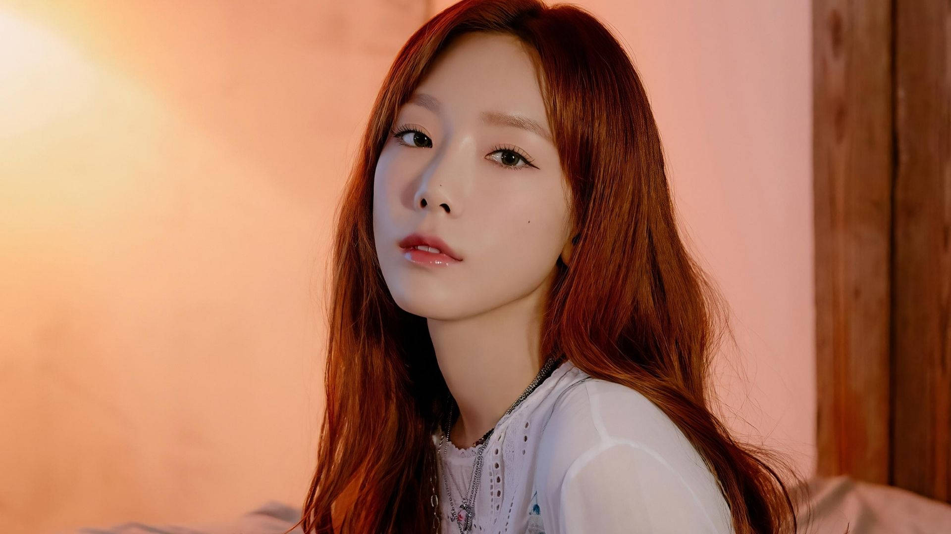 태연 桌布 壁紙 — 一位長紅髮的年輕女性在溫暖的環境燈光下柔和地凝視著相機。