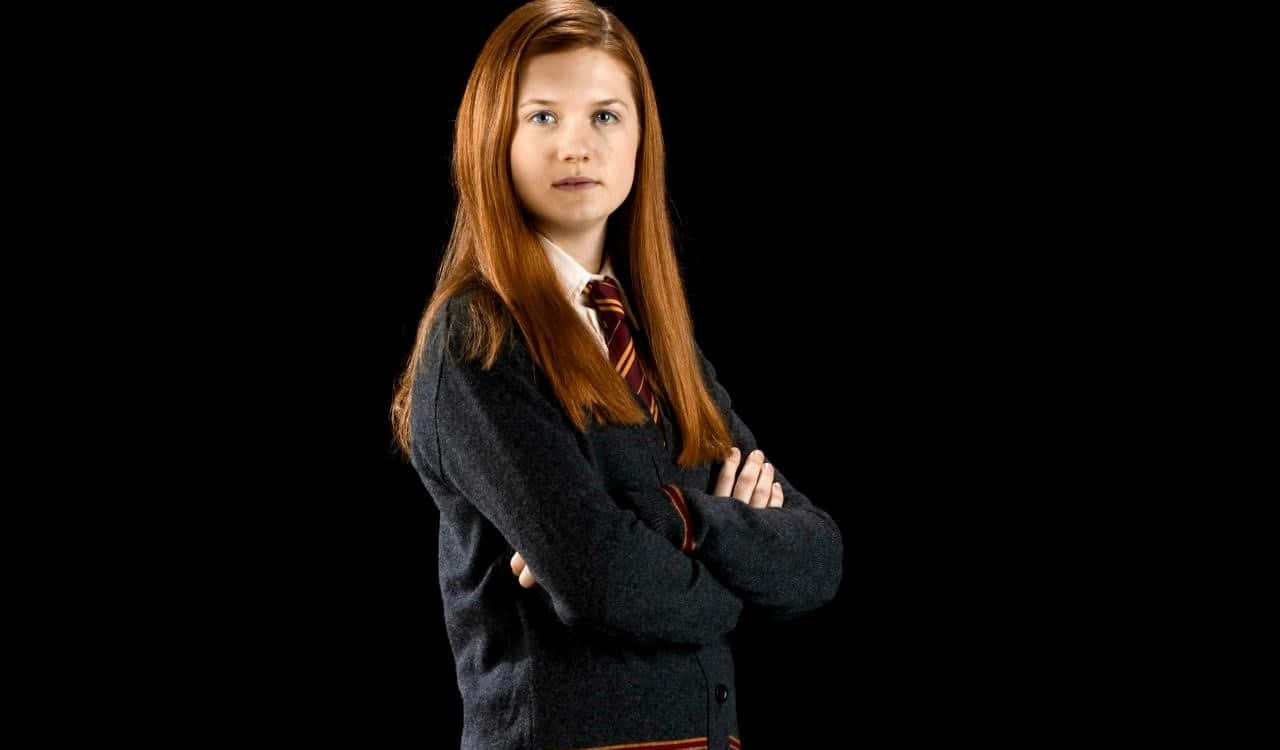 Ginny Weasley 桌布 壁紙 — 一位長紅髮的年輕女性自信地站在黑色背景前，穿著校服，雙臂交叉。