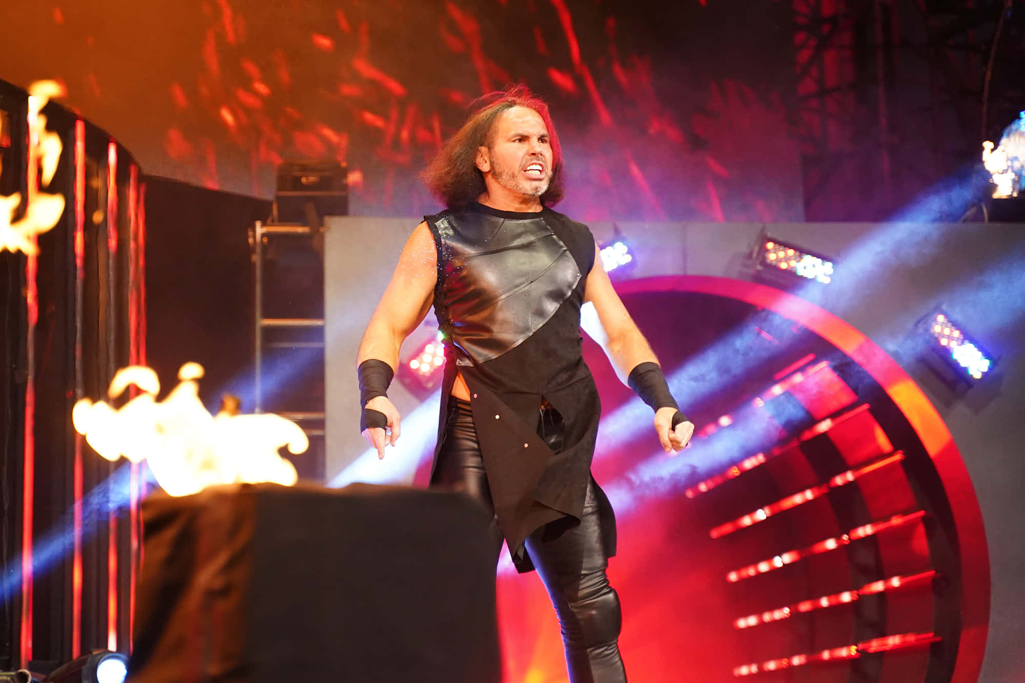 WWE 桌布 壁紙 — 一位長紅髮的摔角手在一個由紅藍燈光照亮、道具噴出火焰的火熱舞台上咆哮。
