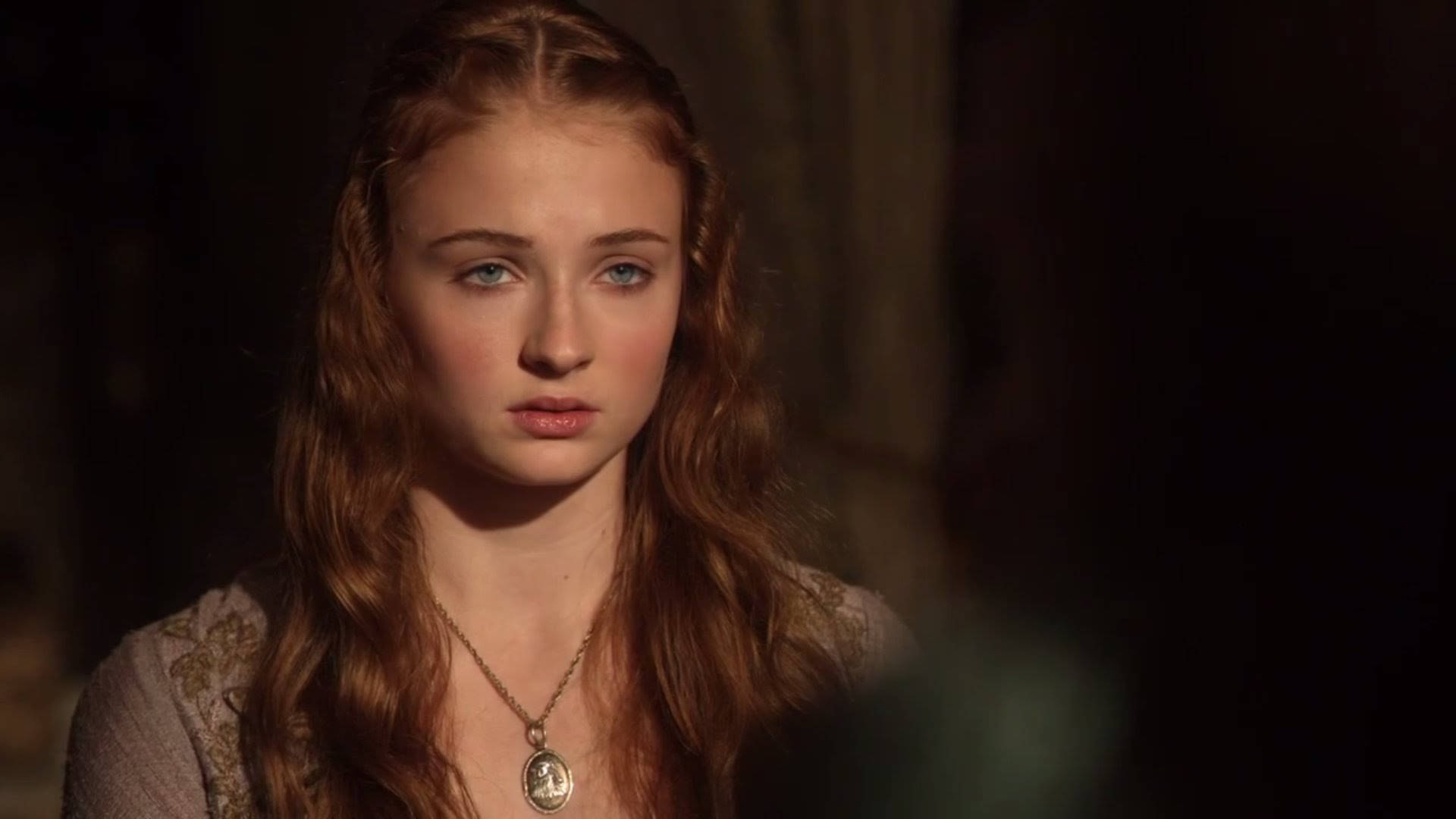 Sansa Stark 桌布 壁紙 — 一位長紅髮藍眼的年輕女性在昏暗的房間裡戴著項鍊，若有所思地凝視著。