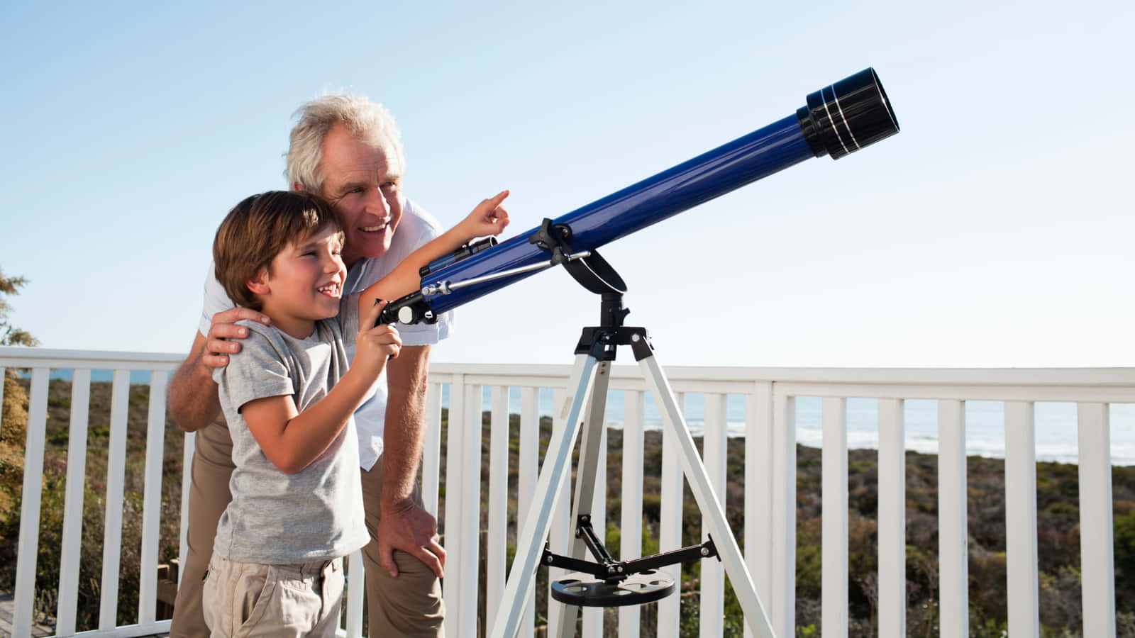 astronomy 桌布 壁紙 — 一位長者和一個小男孩在陽光明媚、俯瞰海洋的陽臺上，透過藍色望遠鏡分享歡樂時刻。