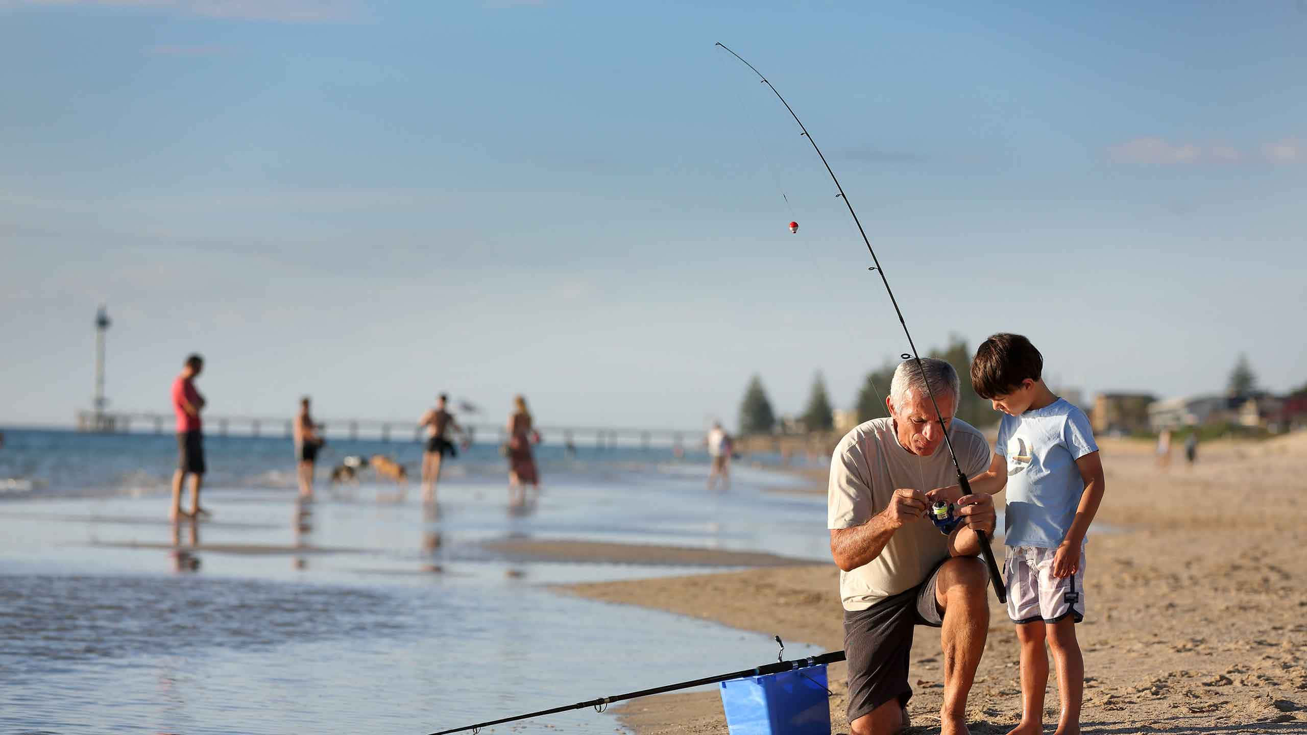 Fishing Rod 桌布 壁紙 — 一位長者和一個小男孩在陽光明媚的海灘上一起釣魚，享受寧靜的時光，海面有輕柔的波浪和遠處的碼頭。