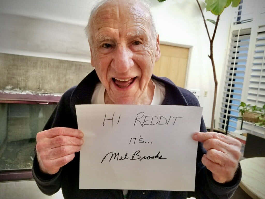 Celebrity Humor Memes 桌布 壁紙 — 一位長者帶著喜悅的表情，在室內手持一張手寫便條，上面寫著「HI REDDIT IT'S... Mel Brooks」。