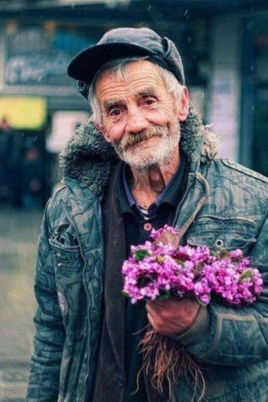 Emotional 桌布 壁紙 — 一位長者帶著溫暖的笑容，在模糊的城市背景下手持一束紫色花朵。