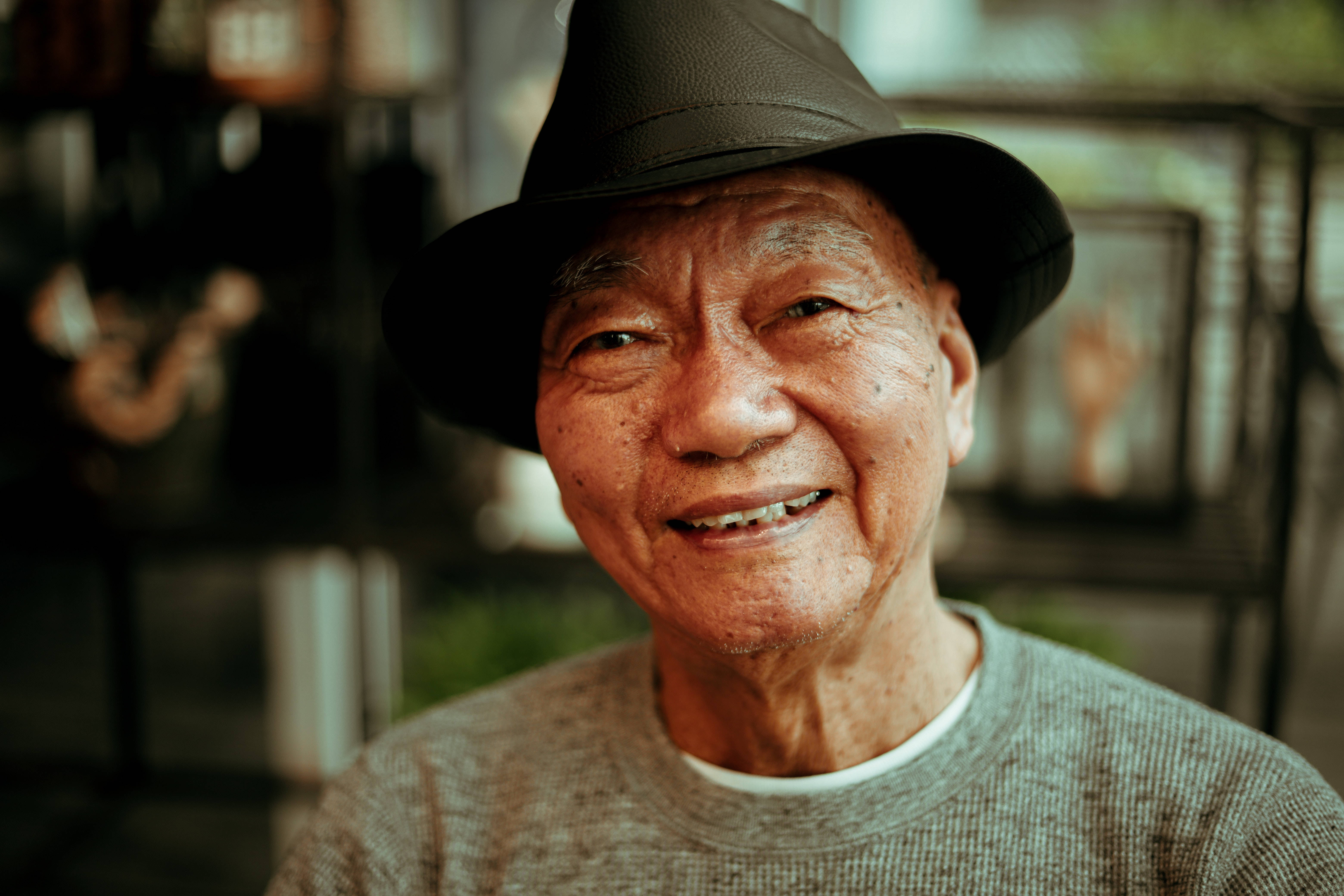 人像動態 桌布 壁紙 — 一位長者帶著溫暖的笑容，戴著黑色軟呢帽，穿著有紋理的毛衣，背景是柔和模糊的室內環境。