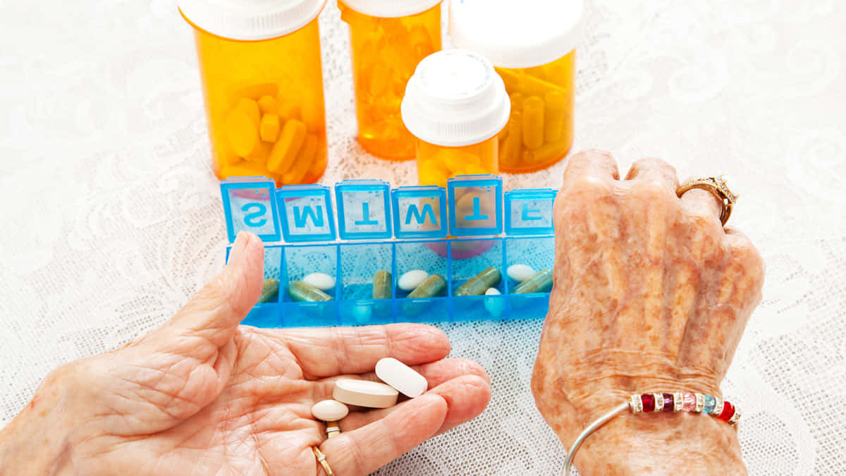pills 桌布 壁紙 — 一位長者的手在蕾絲桌布上，拿著藥丸，旁邊有一個每週藥丸整理器和處方藥瓶。
