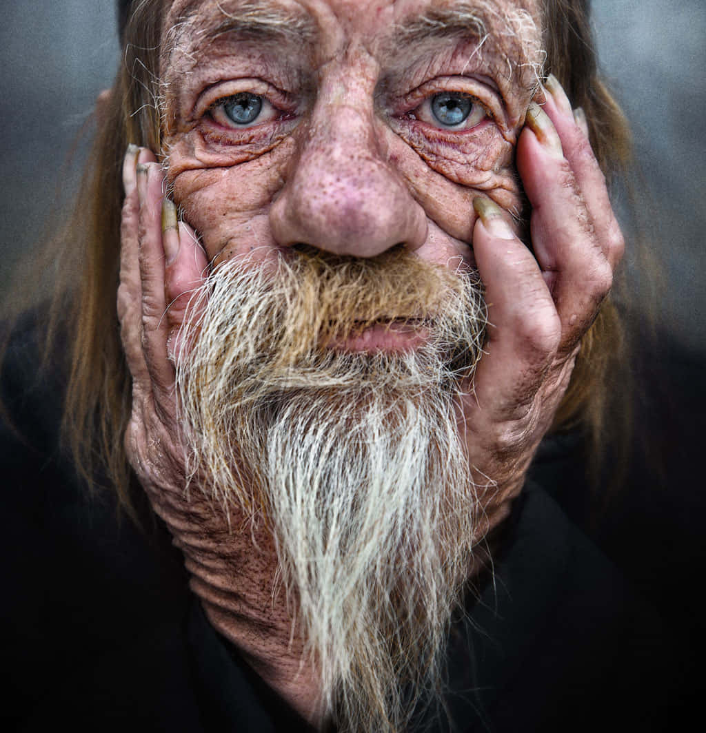 人像動態 桌布 壁紙 — 一位長者的特寫肖像，他有深深的皺紋、藍色眼睛和長長的白鬍子，用粗糙的雙手輕輕托著臉頰。