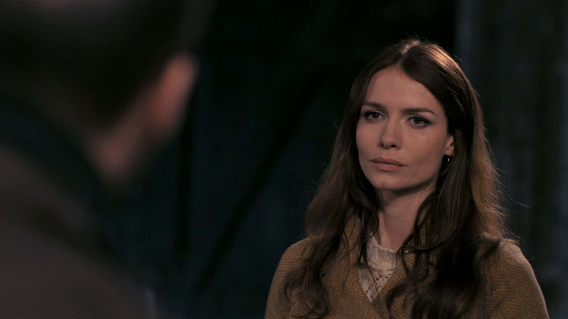 Saffron Burrows 桌布 壁紙 — 一位長著棕色長髮的女性在光線昏暗的室內環境中專注地凝視著一個模糊的人。