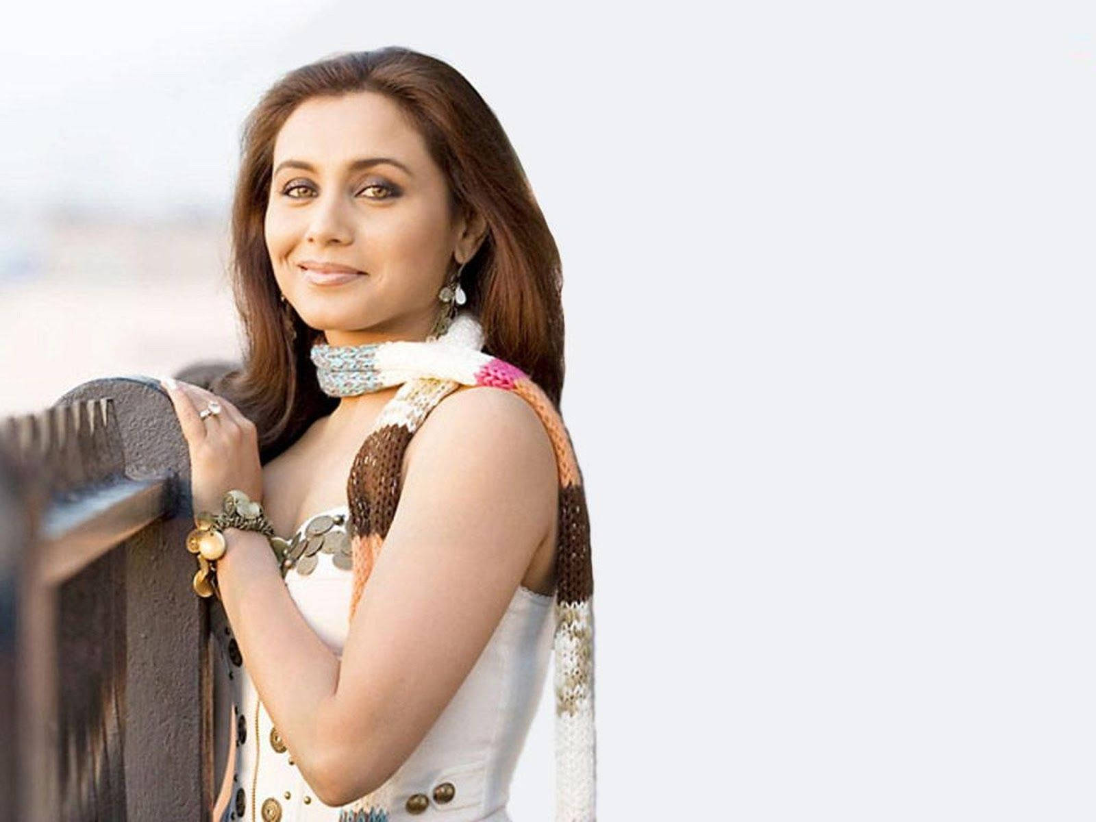 Rani Mukerji 桌布 壁紙 — 一位長著棕色長髮的女性微笑著靠在欄杆上，身穿白色連衣裙和彩色針織圍巾。