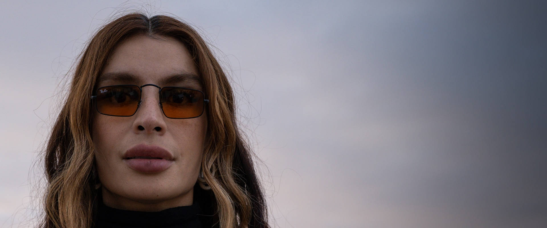 Sunglasses 桌布 壁紙 — 一位長著棕色長髮的女性戴著琥珀色太陽眼鏡，背景是陰沉的陰天。