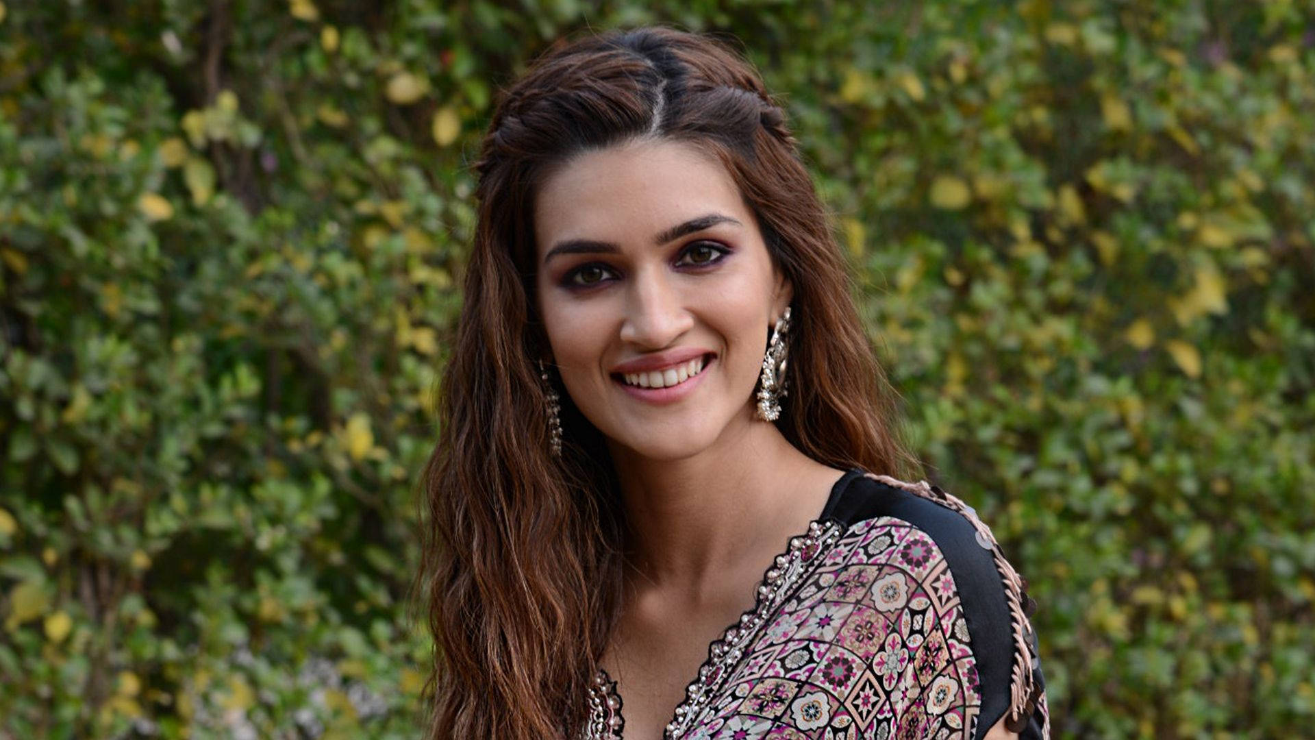 Kriti 桌布 壁紙 — 一位長著棕色長髮的容光煥發的女性，在茂密的綠色樹葉背景下溫暖地微笑。