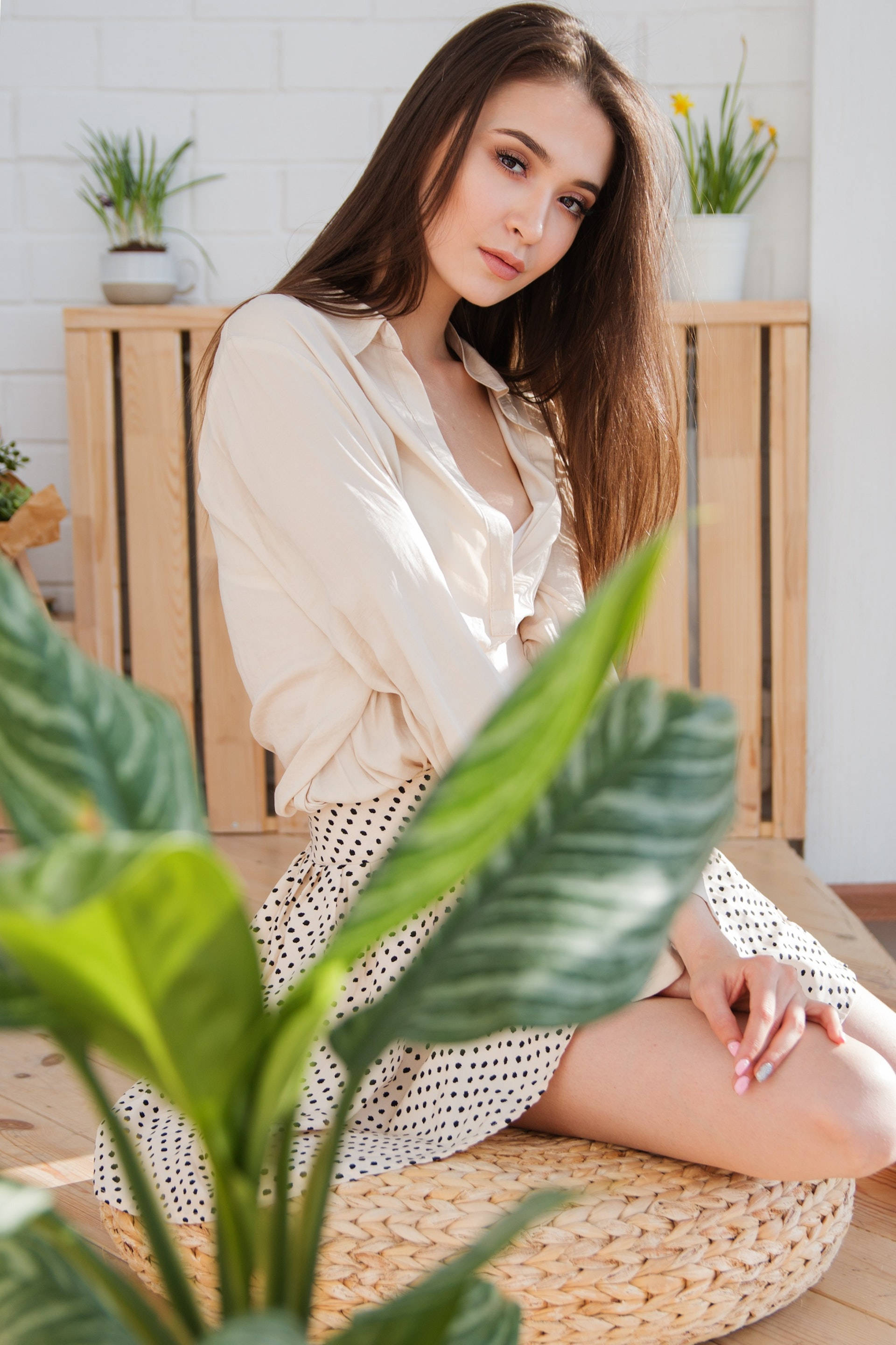 lovely girl 桌布 壁紙 — 一位長著棕色長髮的年輕女子平靜地坐在編織墊上，周圍是茂盛的綠色植物，身處明亮的簡約房間。
