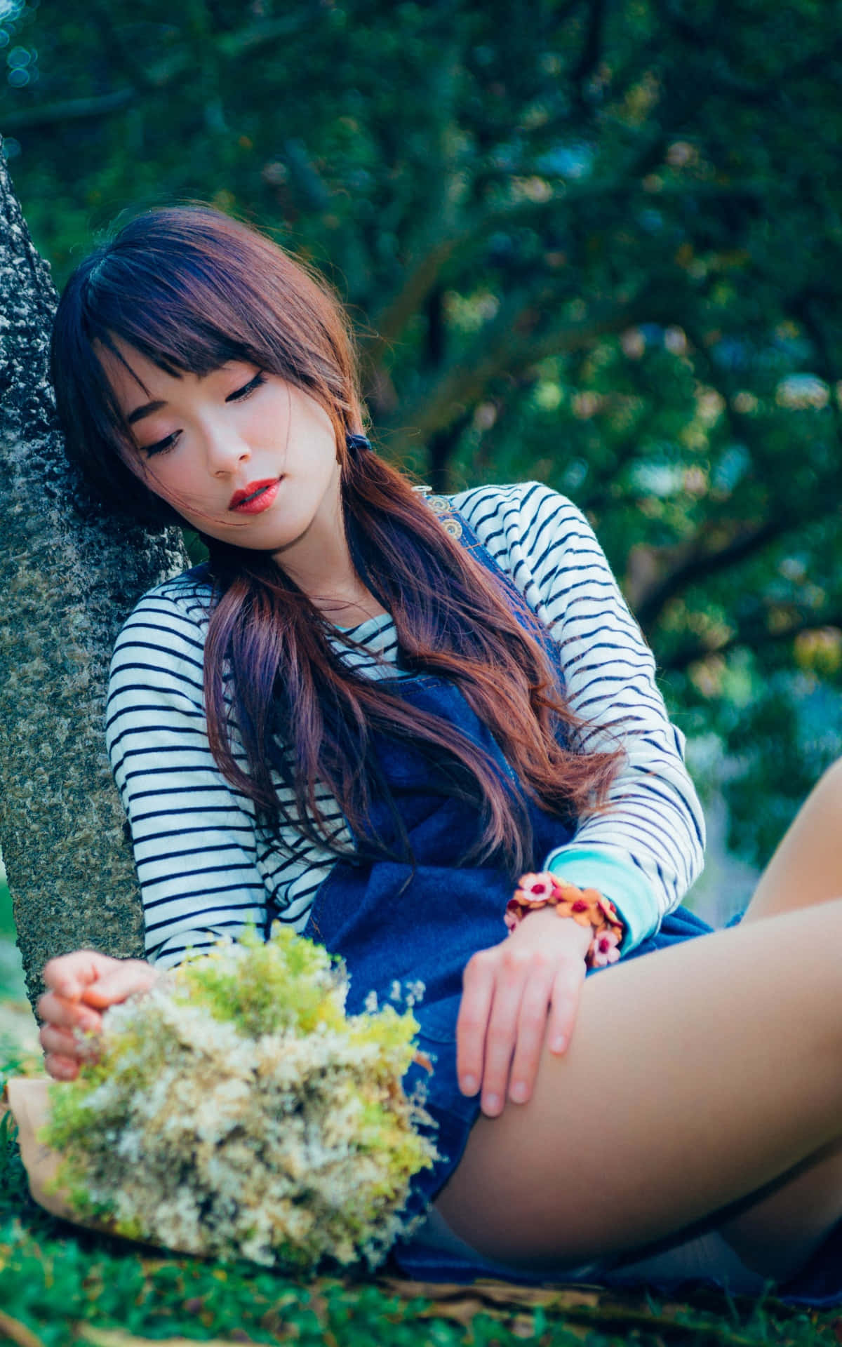 Sitting Alone 桌布 壁紙 — 一位長著棕色長髮的年輕女子靠著樹坐著，在茂密的綠色森林中手持一束苔蘚花束。