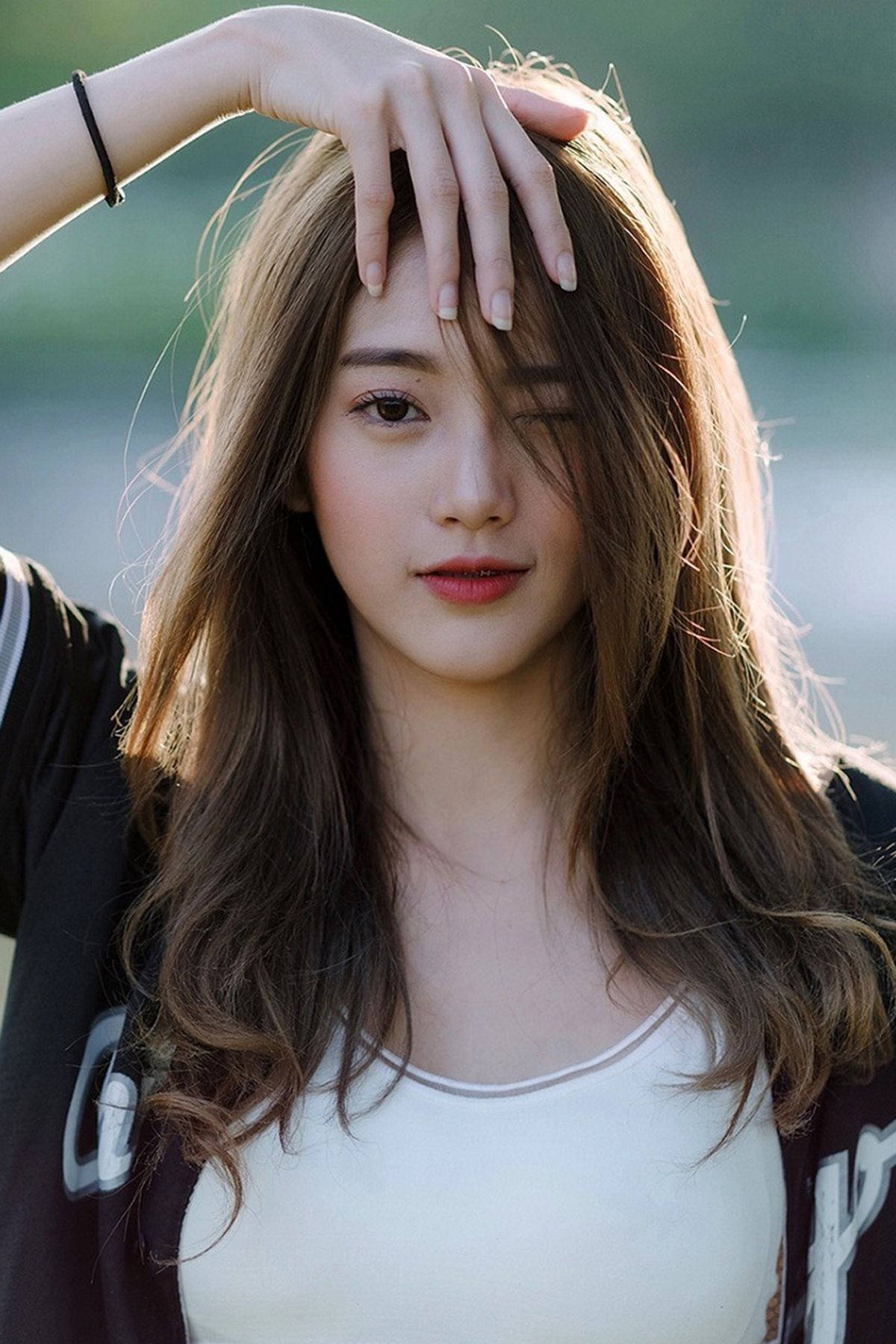 girls 桌布 壁紙 — 一位長著棕色長髮的年輕女性，在柔和的陽光下，以模糊的戶外背景為襯托，輕輕觸碰額頭。