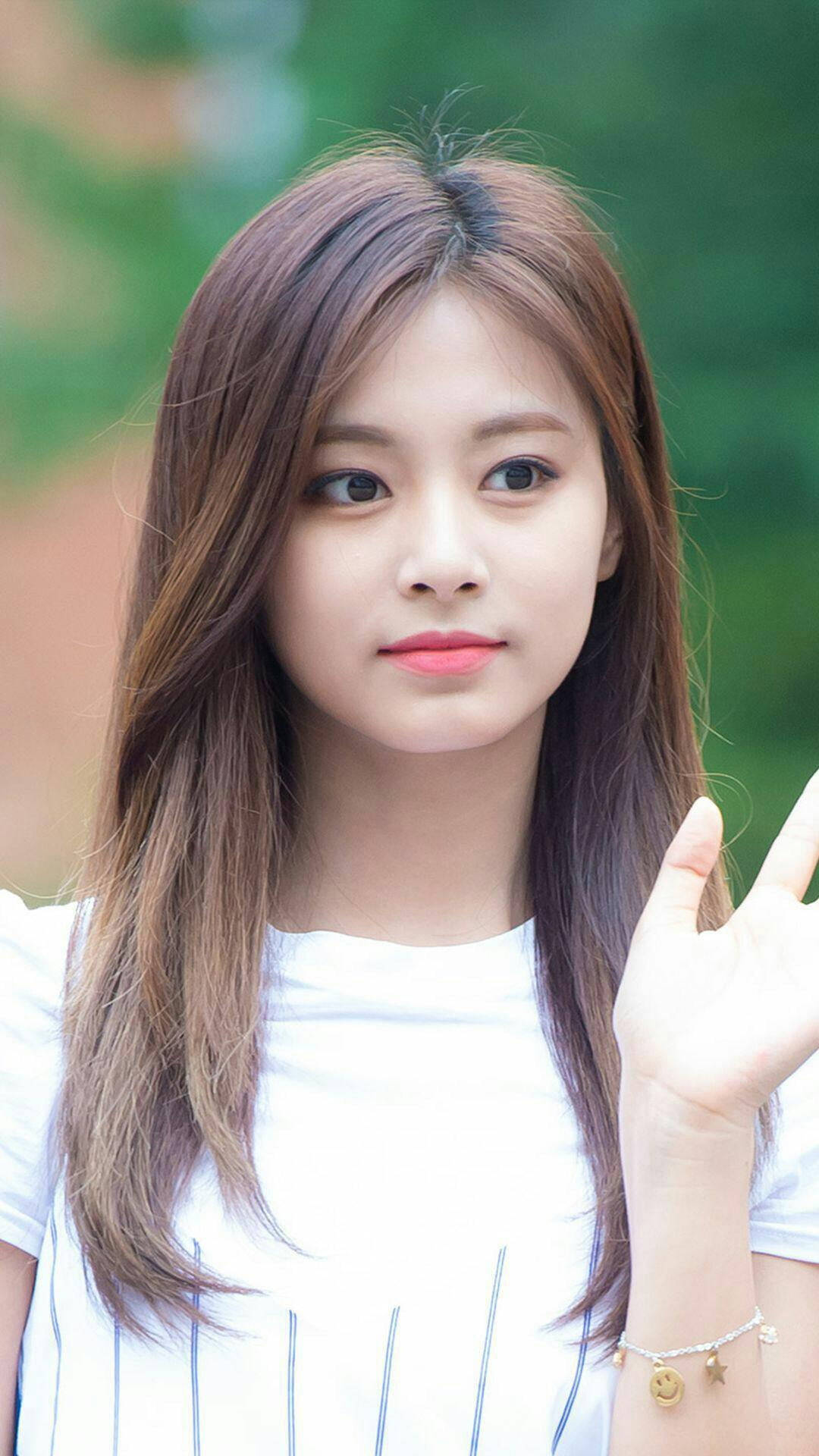 Twice Mina 桌布 壁紙 — 一位長著棕色長髮的年輕女性，在模糊的綠色背景前微笑著揮手。