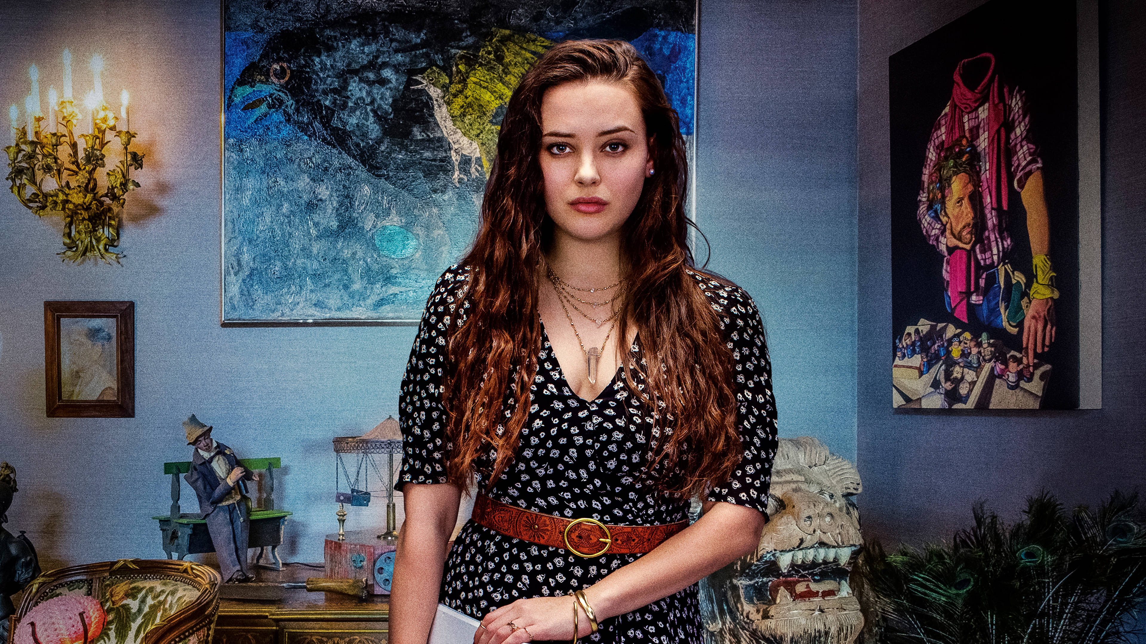 Katherine Langford 桌布 壁紙 — 一位長著棕色長髮的年輕女性站在一個藍色牆壁的房間裡，房間裡裝飾著不拘一格的藝術品和復古裝飾，在溫暖的吊燈燈光下。