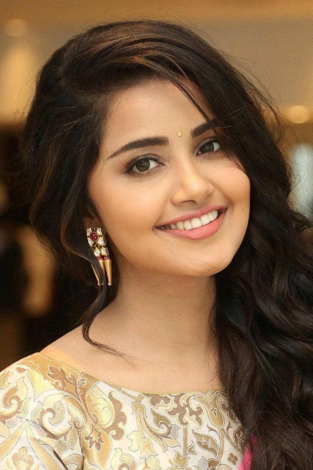 anupama parameswaran 桌布 壁紙 — 一位長著深色波浪長髮的容光煥發的女性，戴著華麗的金耳環和圖案襯衫，溫暖地微笑。