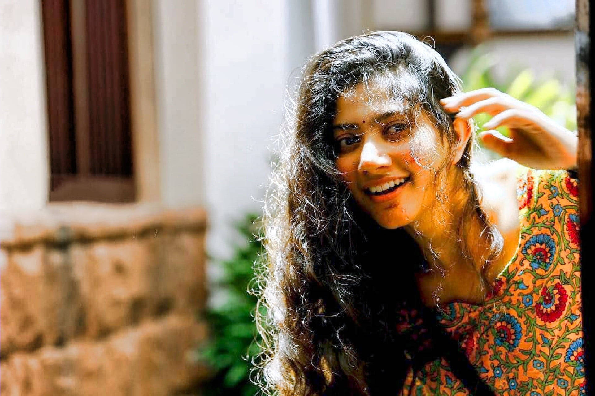 Sai Pallavi 桌布 壁紙 — 一位長著深色長捲髮的快樂女性，在陽光照射的石建築附近溫暖地微笑，同時觸摸她的頭髮。