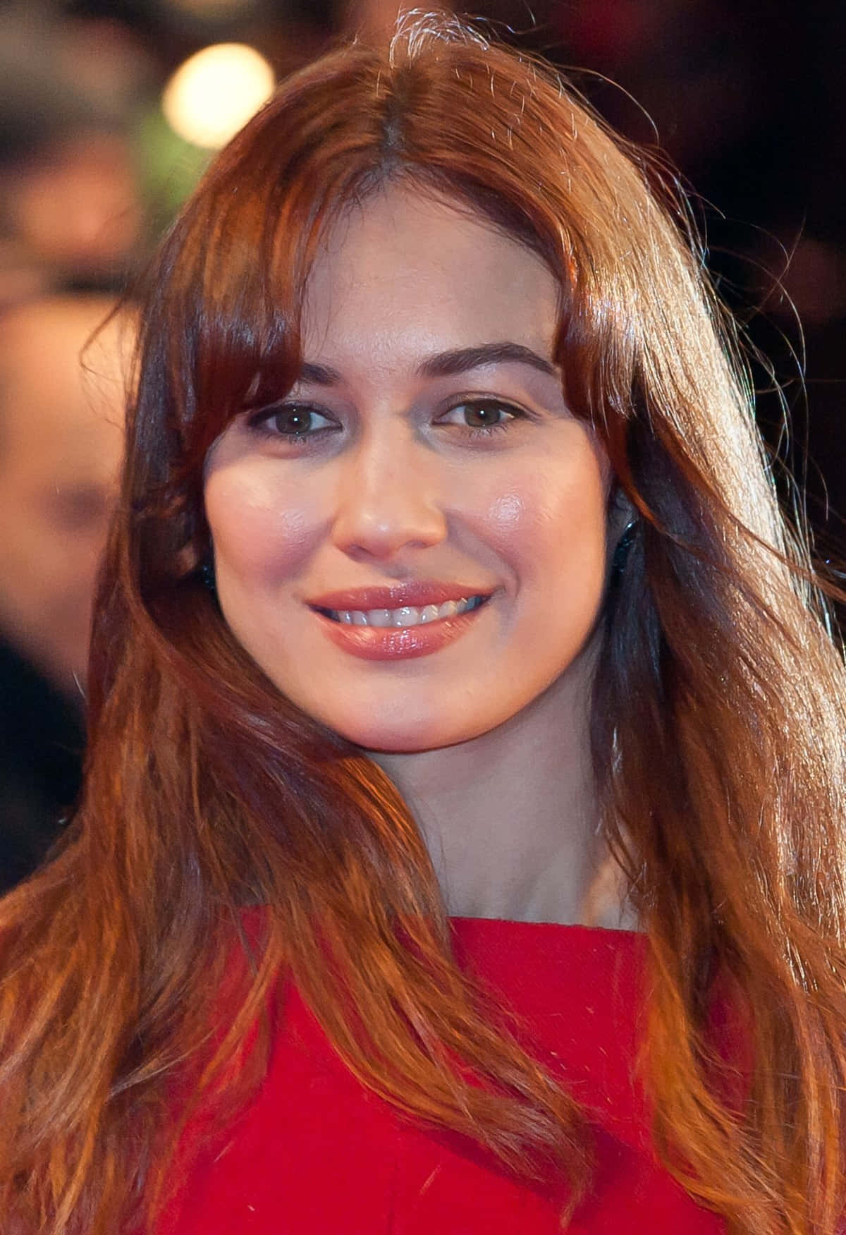 Olga Kurylenko 桌布 壁紙 — 一位長著赤褐色長髮的女性穿著鮮紅色上衣溫暖微笑的特寫肖像。