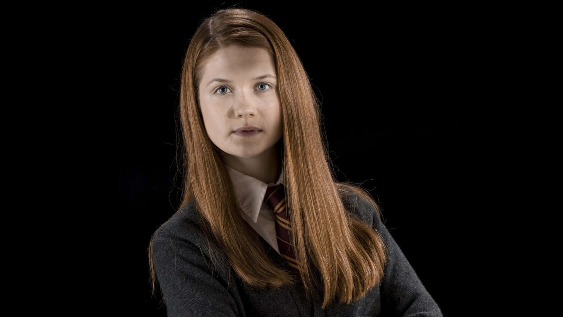 Ginny Weasley 桌布 壁紙 — 一位長著赤褐色長髮的年輕女性，在黑色背景下穿著深色校服和條紋領帶。