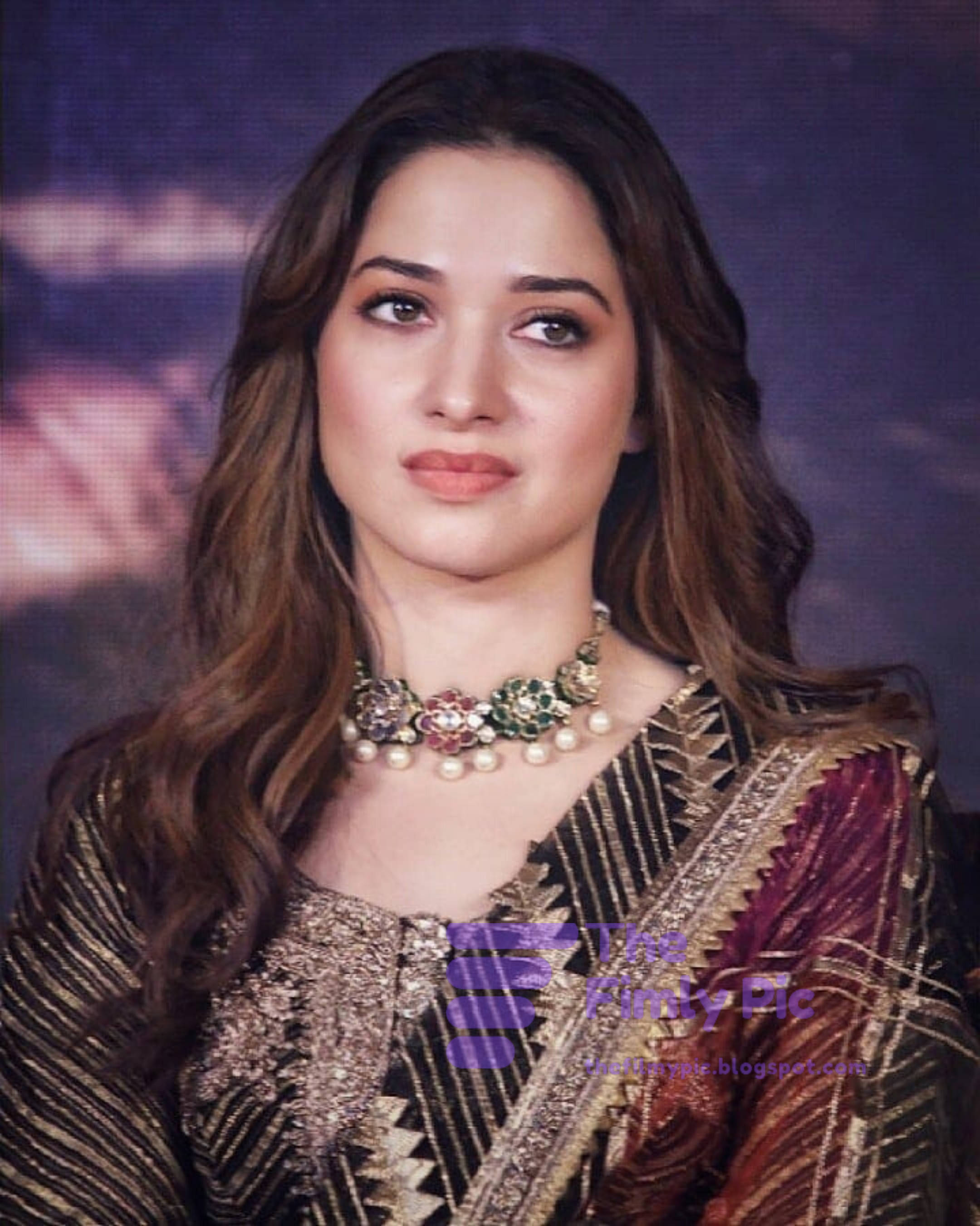 tamannaah bhatia 桌布 壁紙 — 一位長著長波浪棕色頭髮的女性，穿著華麗的金色和栗色刺繡服裝，戴著花卉珍珠項鍊，背景是模糊的紫色。