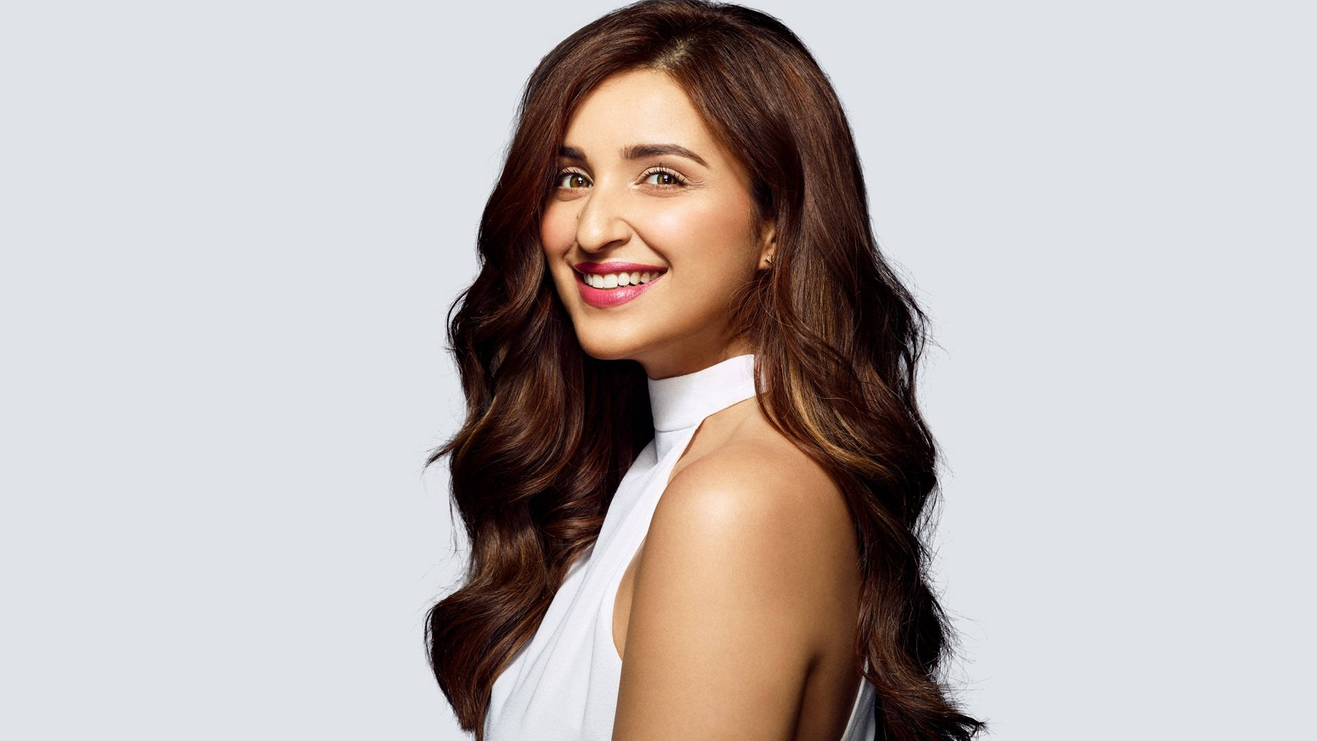 Parineeti 桌布 壁紙 — 一位長著長波浪棕色頭髮的容光煥發的女性，在柔和的灰色背景前穿著白色掛頸上衣，溫暖地微笑。