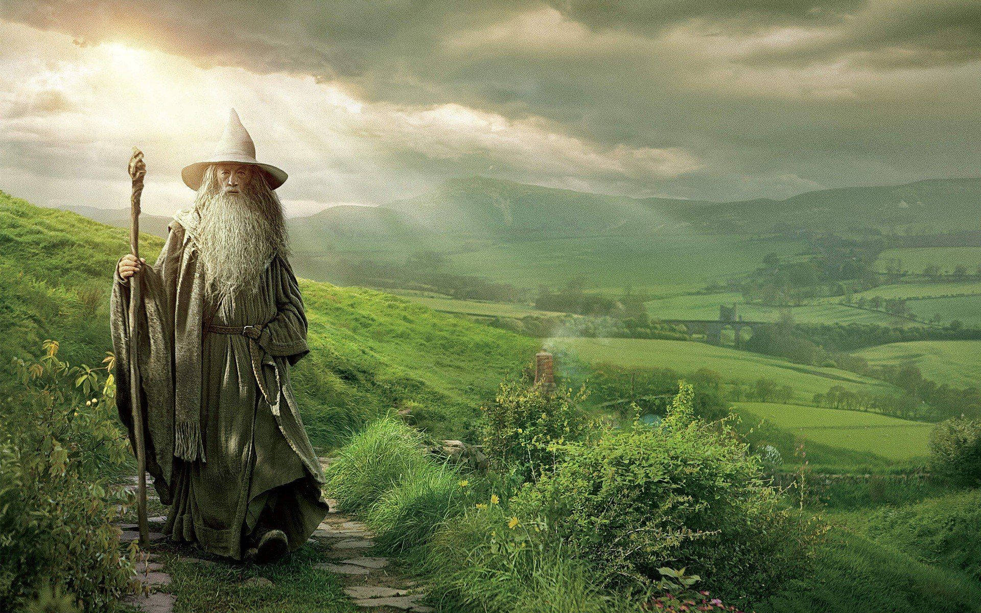 Gandalf 桌布 壁紙 — 一位長著長長白鬍鬚、戴著尖帽子的老巫師站在一條石徑上，身處翠綠的山谷，頭頂是戲劇性的多雲天空。