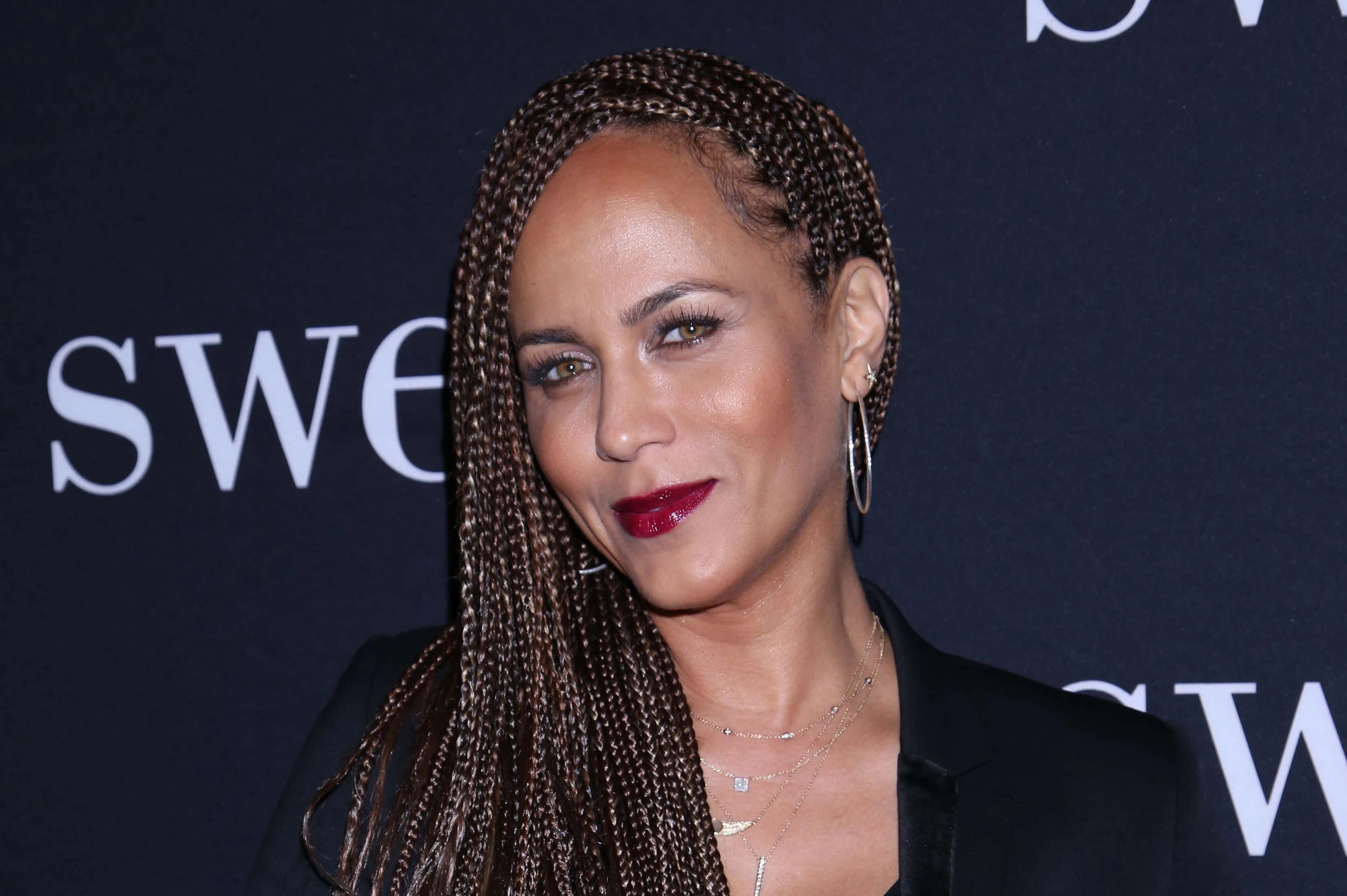 Nicole Ari Parker 桌布 壁紙 — 一位長辮子、紅唇大膽的優雅女性，在深色品牌背景前對著鏡頭微笑。