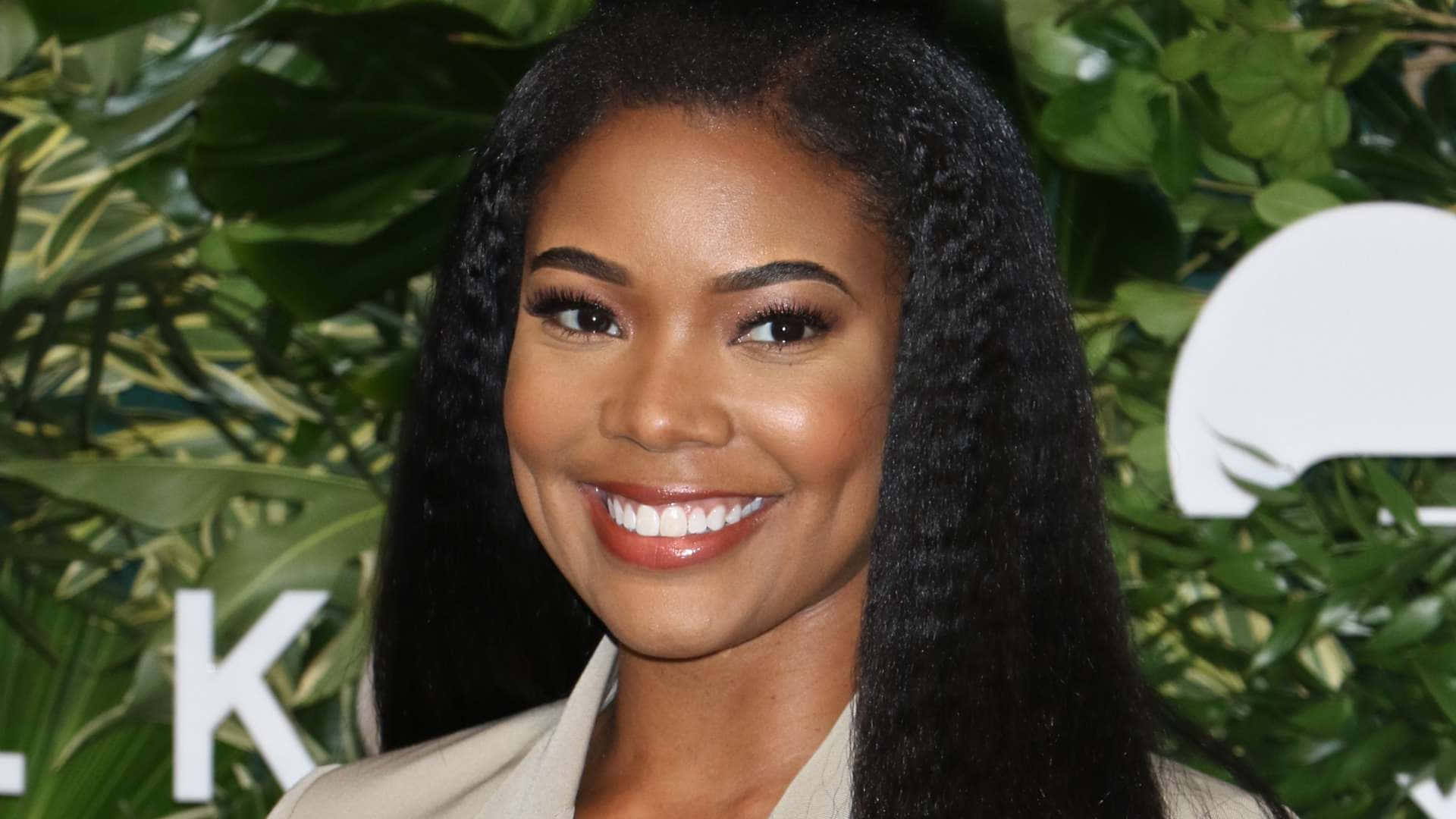 gabrielle union 桌布 壁紙 — 一位長辮子的光彩照人的女性，在茂密的綠色樹葉背景下燦爛微笑。