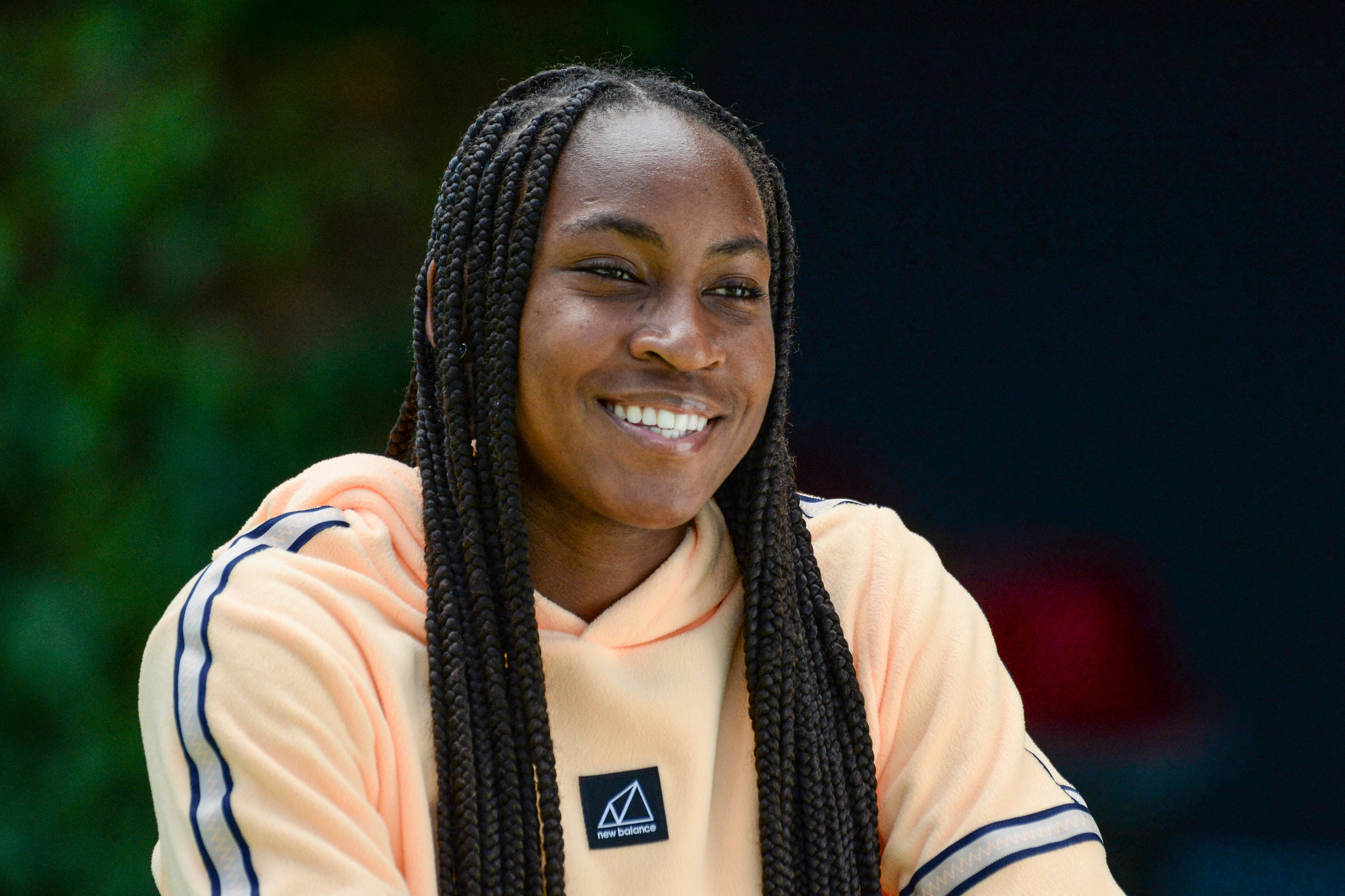 Coco Gauff 桌布 壁紙 — 一位長辮子的快樂女性，穿著帶有藍色條紋的蜜桃色連帽衫，溫暖地微笑。