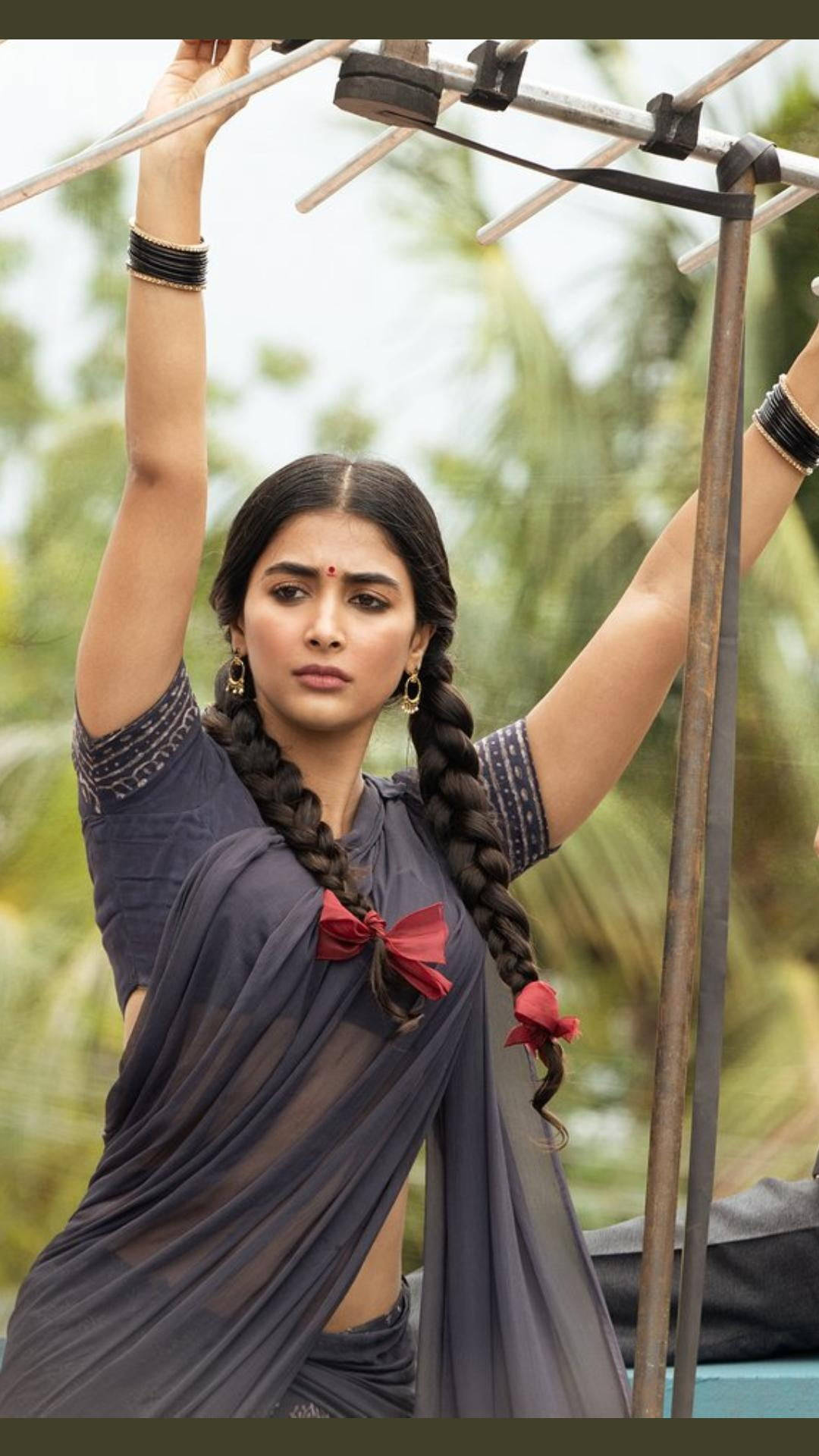 telugu 桌布 壁紙 — 一位長辮子飾有紅色絲帶的女性，在模糊的綠色棕櫚樹背景下向上伸展，同時抓住金屬桿。