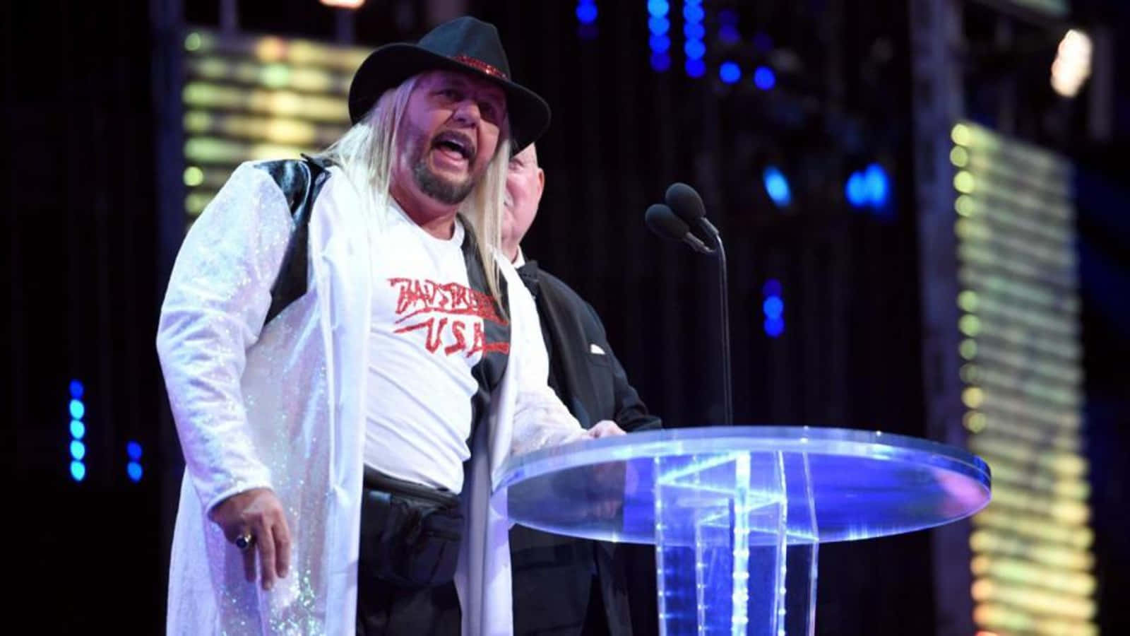 WWE 桌布 壁紙 — 一位長金髮、戴黑帽的摔角手在舞台燈光下，於發光的藍色講台上熱情演講。