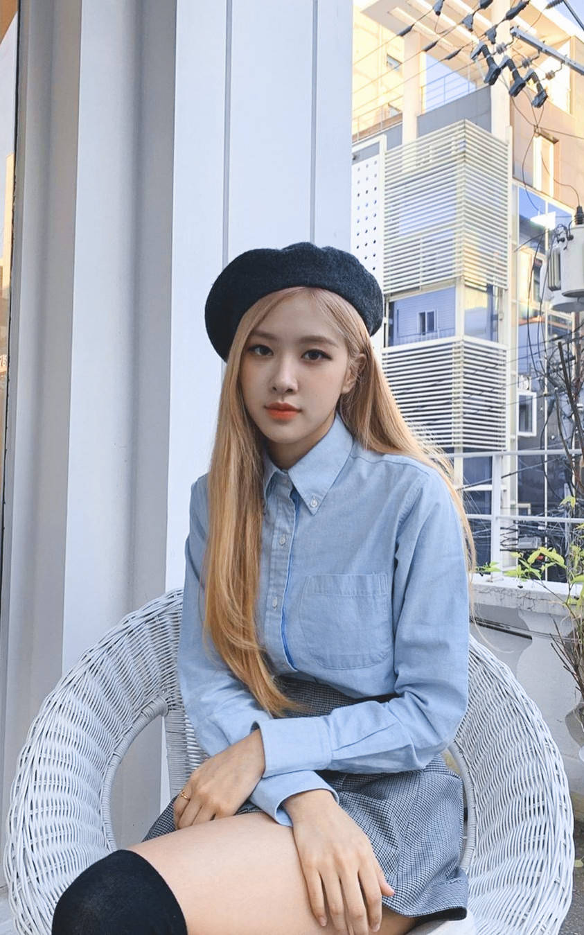 Blackpink Aesthetic 桌布 壁紙 — 一位長金髮、戴黑色貝雷帽的年輕女性，在現代都市背景下，靜靜地坐在白色藤椅上。