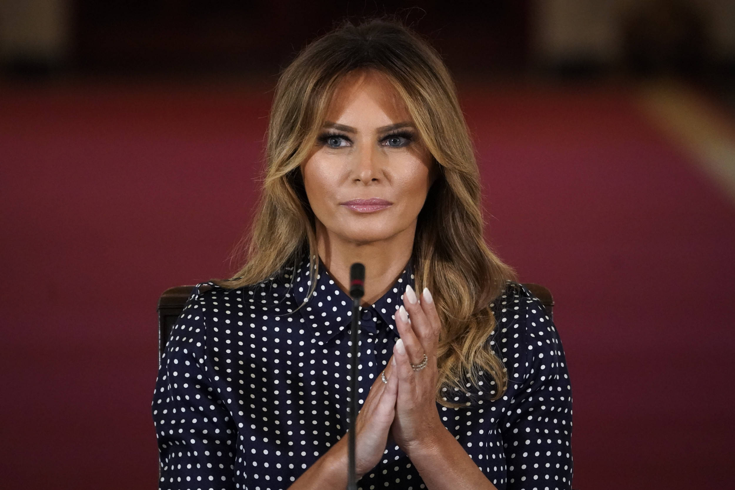 Melania 桌布 壁紙 — 一位長金髮、身穿圓點襯衫的女性坐在講台上，對著深紅色背景拍手。