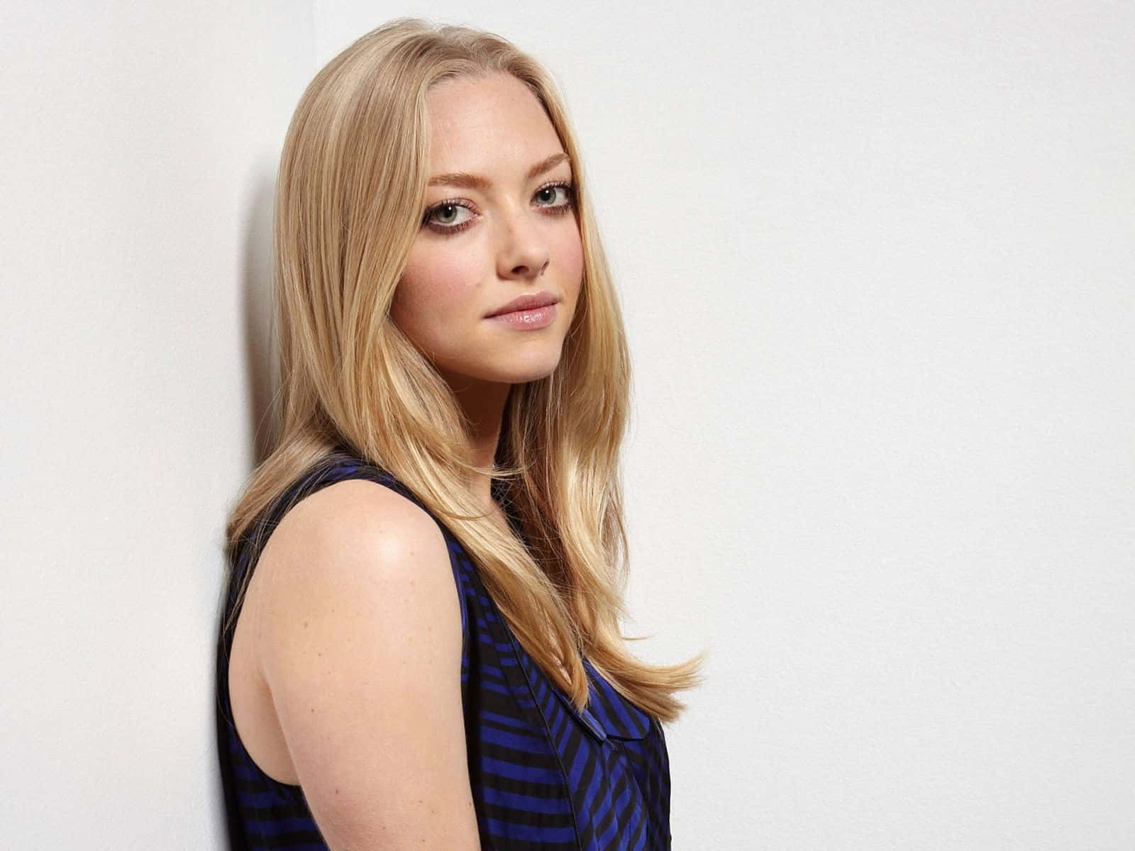 Amanda Seyfried 桌布 壁紙 — 一位長金髮、身穿藍色條紋上衣的女性，在柔和的工作室燈光下靠著純白牆壁。