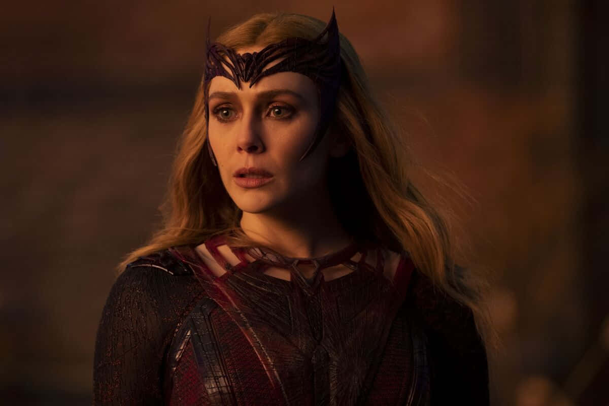Marvel Profile 桌布 壁紙 — 一位長金髮的女性，在溫暖昏暗的燈光下，戴著深色華麗的頭飾和紅色質感服裝。